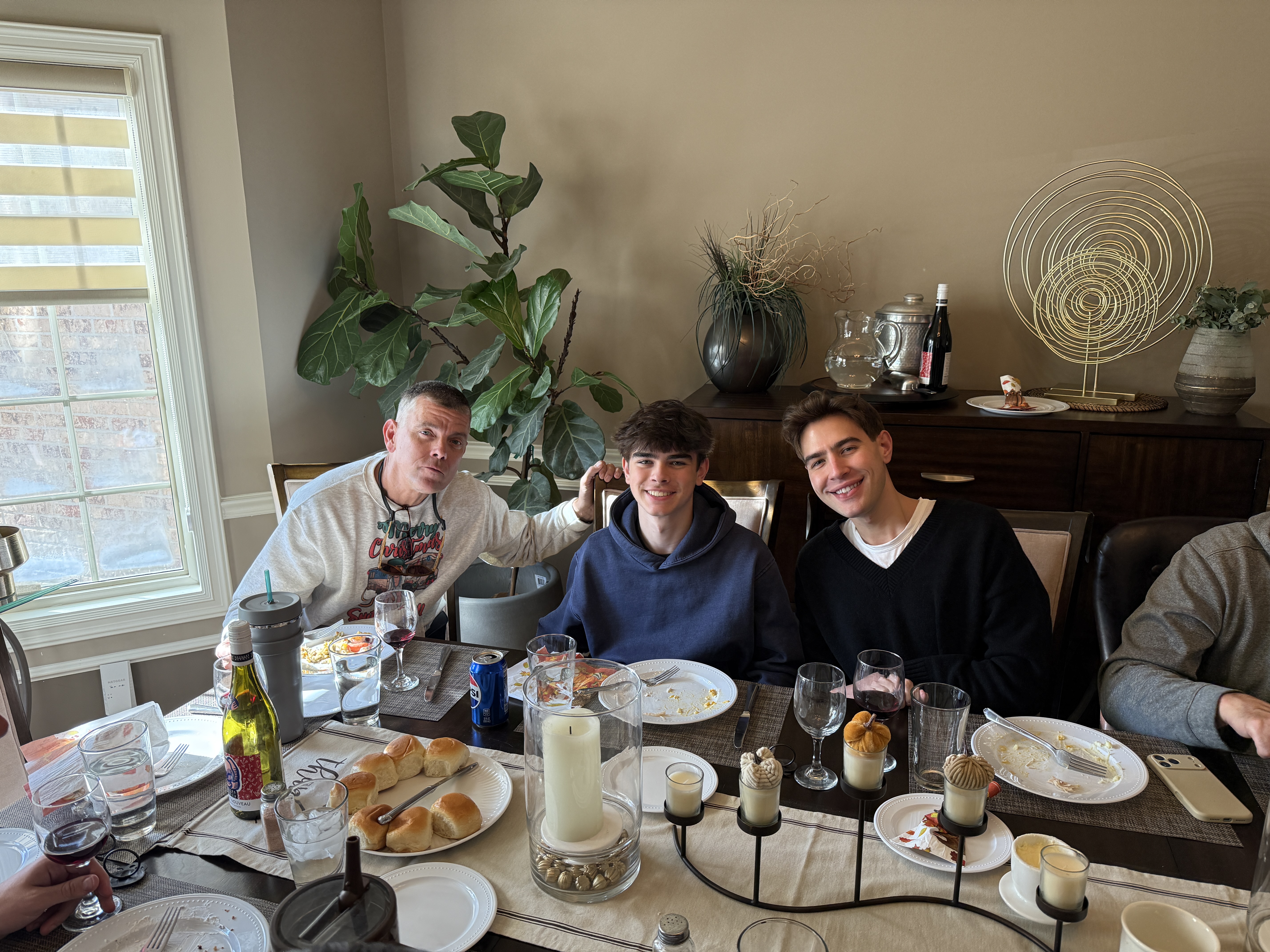 Three people enjoying a meal together at a dining table, with plates and drinks, surrounded by a cozy interior and plants.