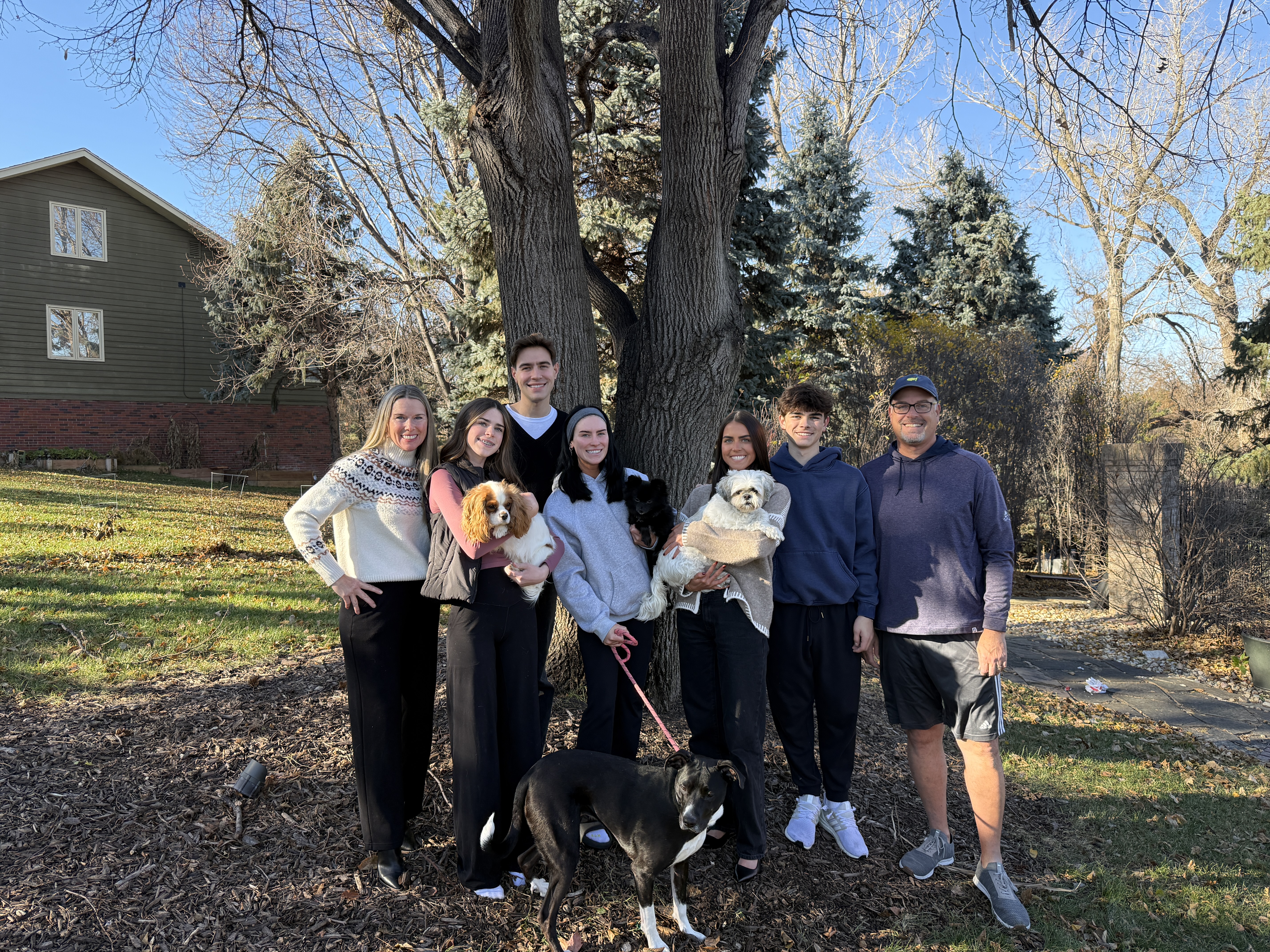 A group of seven people and three dogs pose together outdoors, standing in front of a large tree and a house. The scene shows a sunny day with clear skies and trees in the background.