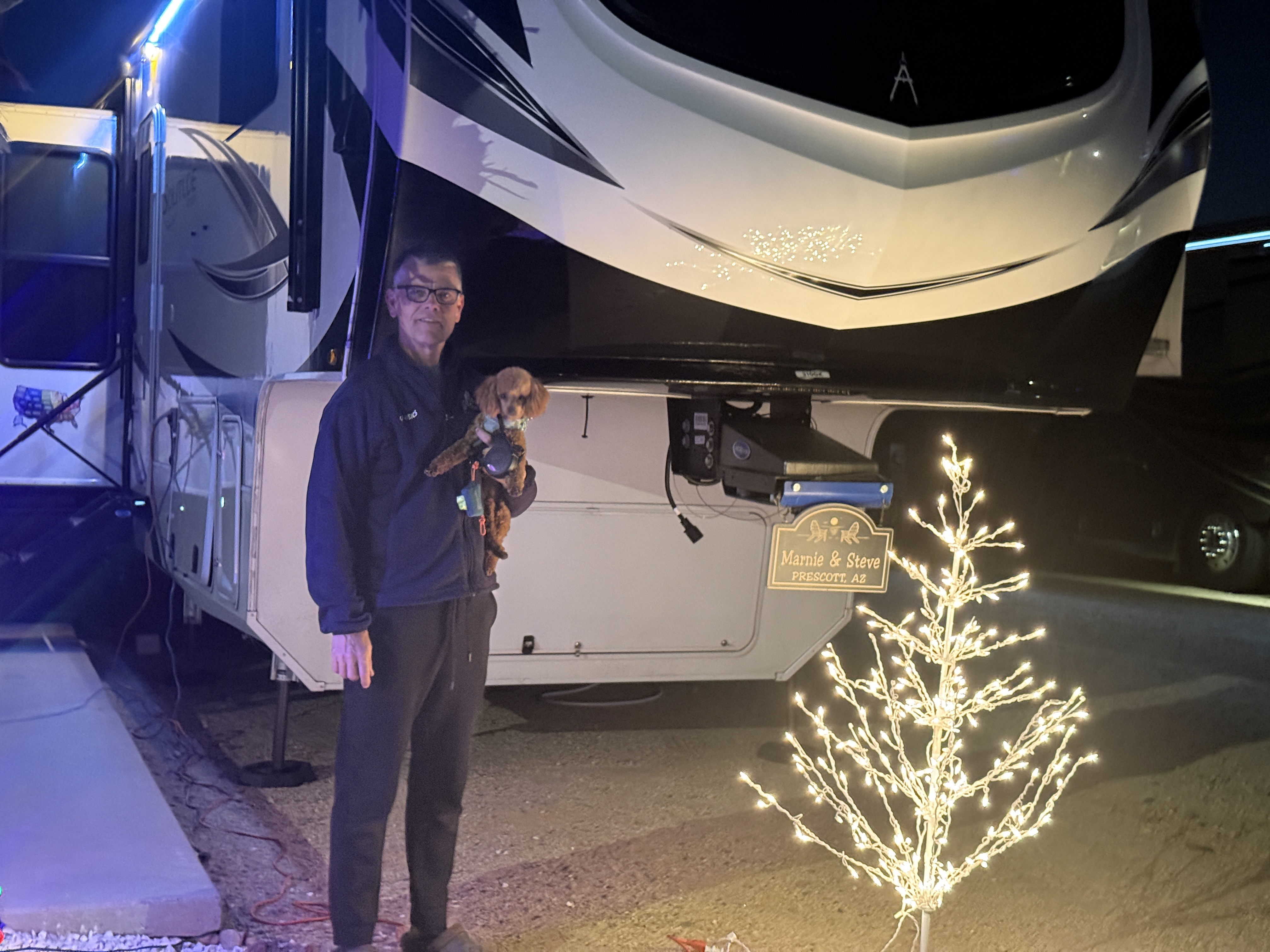 A man with glasses holds a small dog while standing in front of a recreational vehicle at night, with a lit decorative tree nearby.