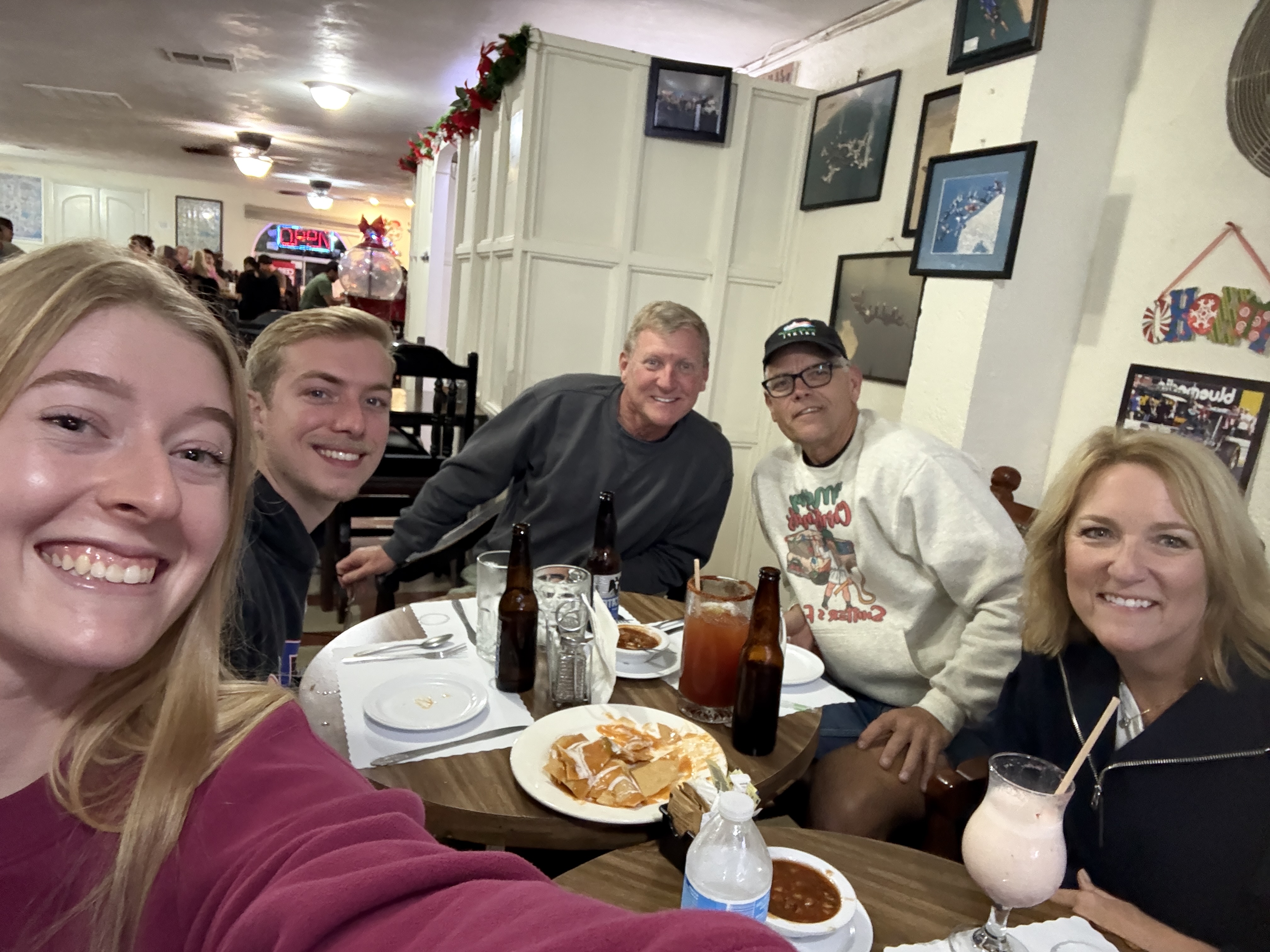 A group of five people smiling together in a restaurant setting, enjoying food and drinks at a table.