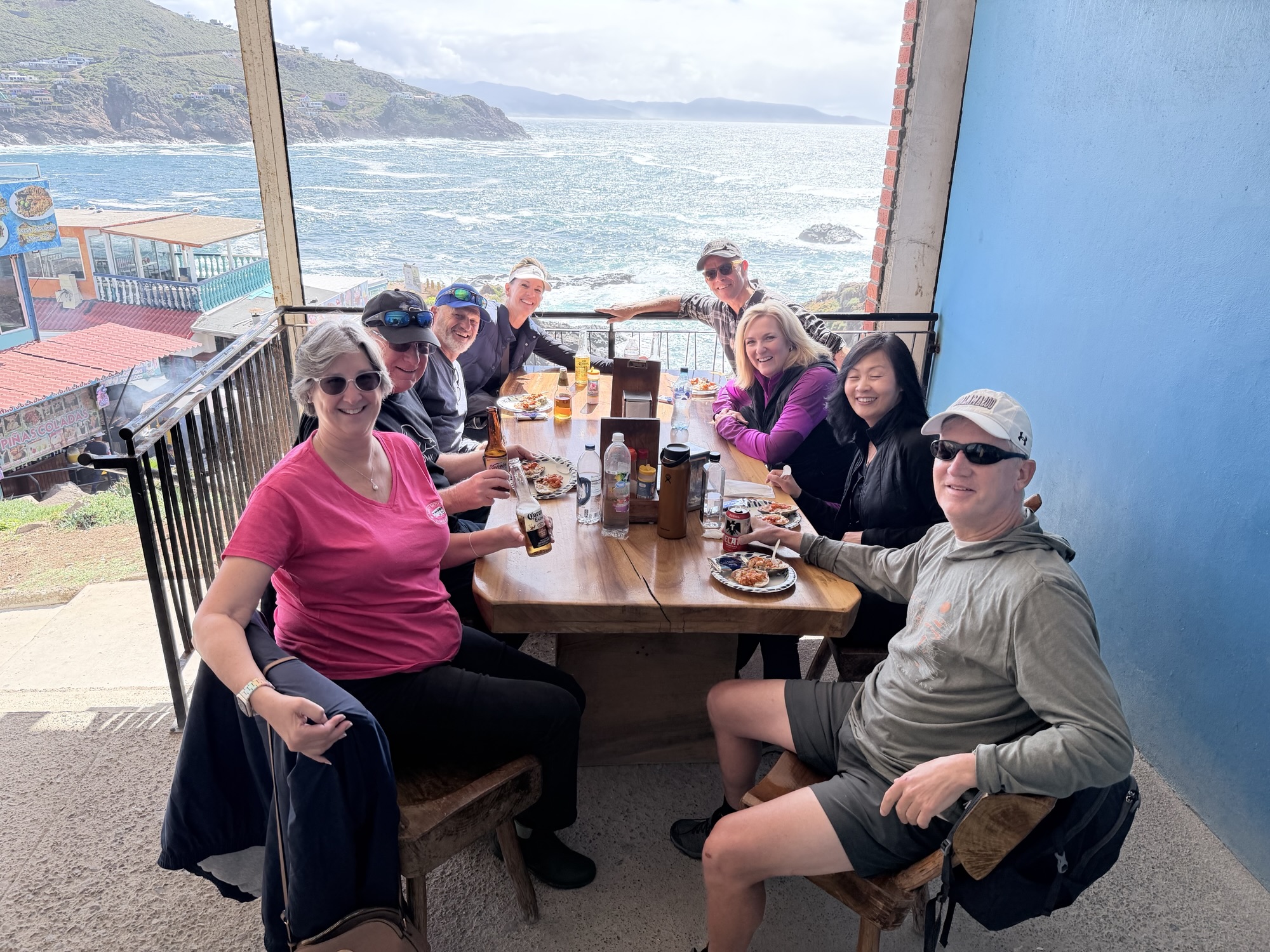 Group of eight people sitting around a wooden table enjoying food and drinks at a coastal restaurant with a view of the ocean.