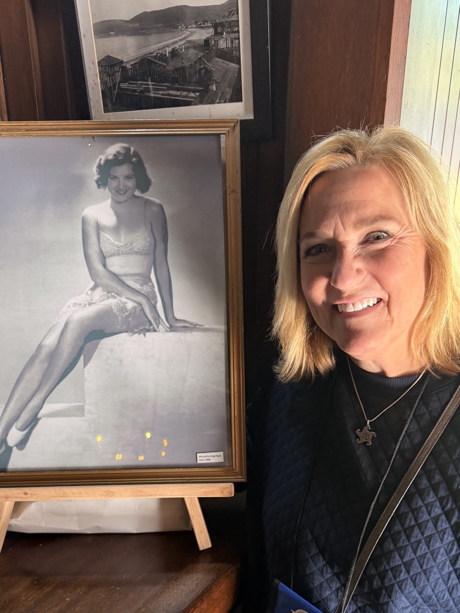 A woman smiling next to a framed black and white photo of a glamorous woman in vintage clothing, with a background featuring additional framed images.