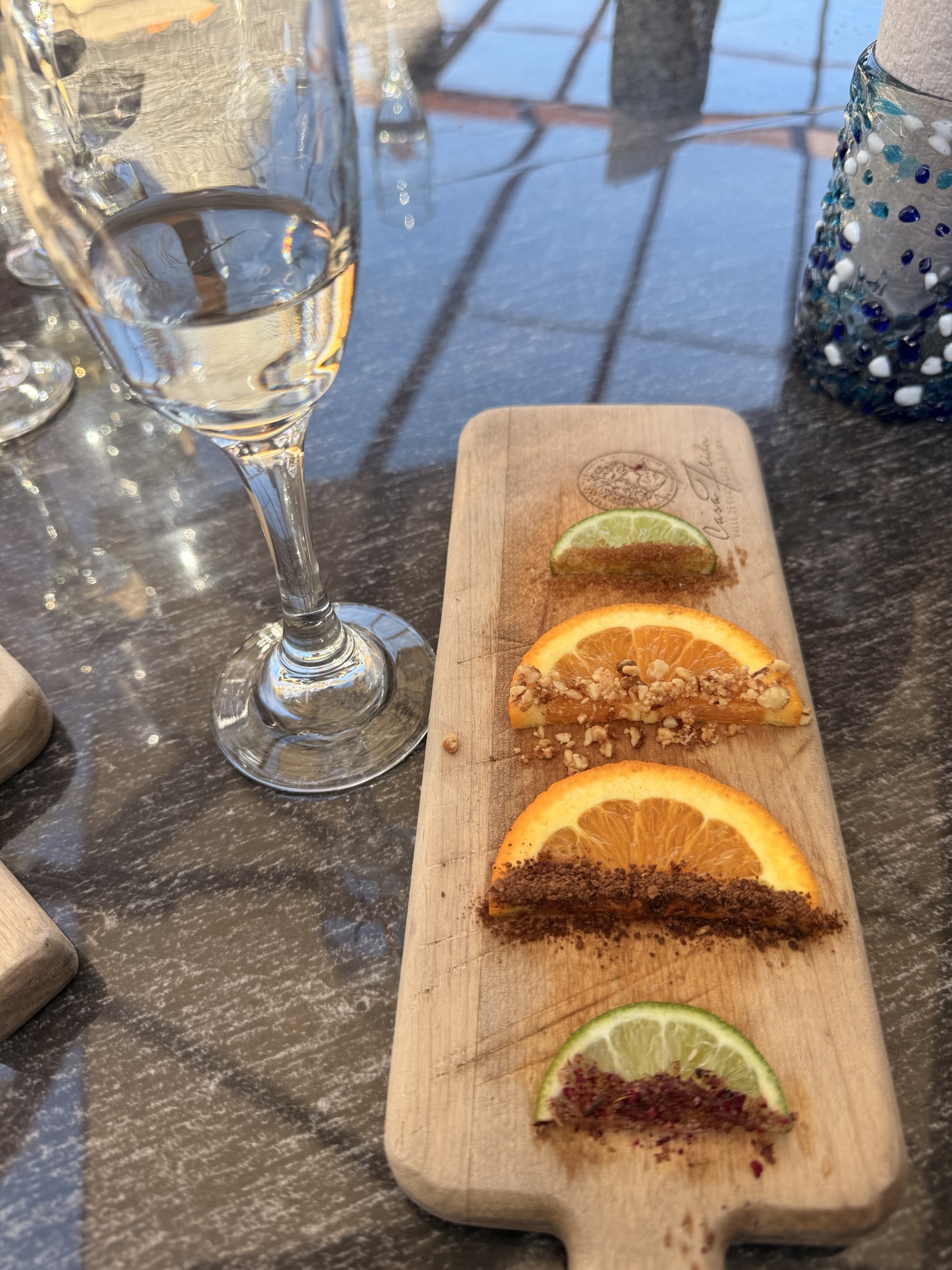A wooden serving board with slices of orange and lime topped with different seasonings, accompanied by a glass of clear liquid, reflecting light.