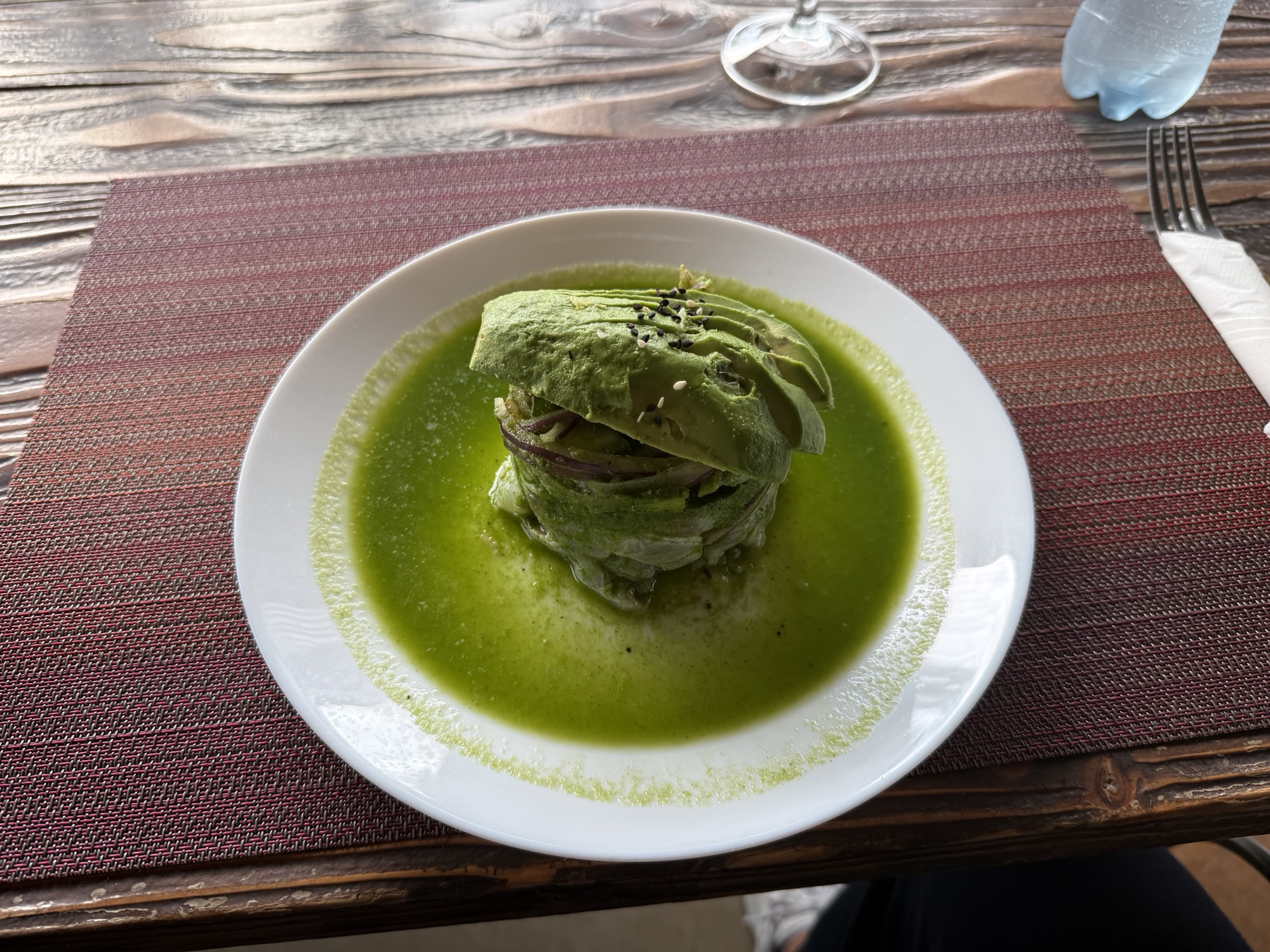 A plated dish featuring a stack of green avocado slices layered with onions, served in a vibrant green sauce, on a textured placemat.