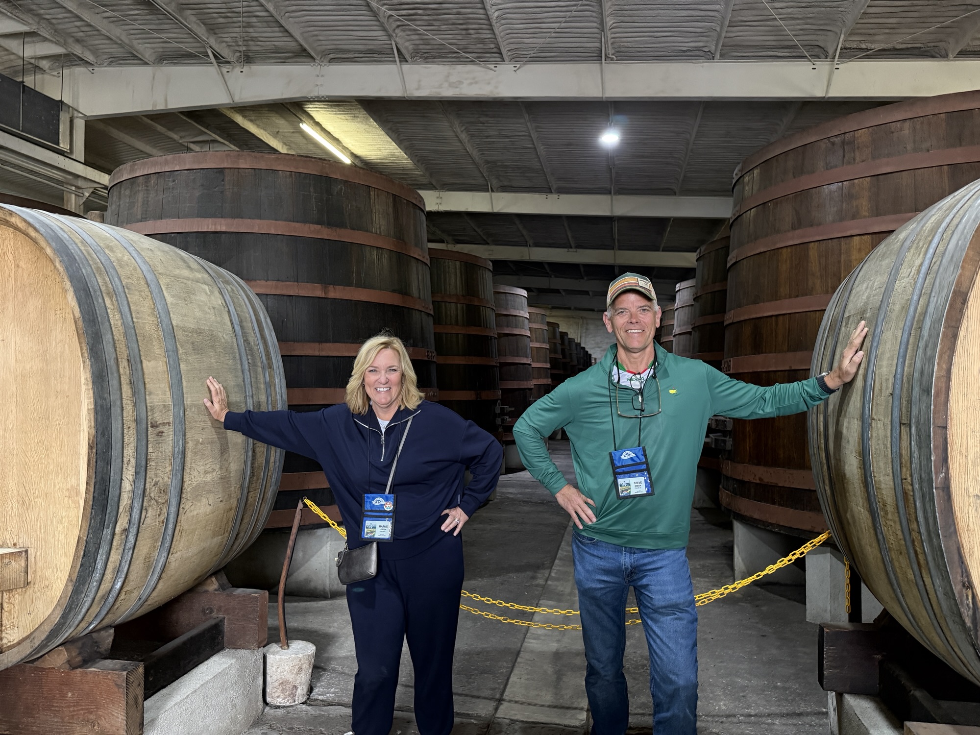 Two smiling people standing beside large wooden barrels in a winery or brewery.