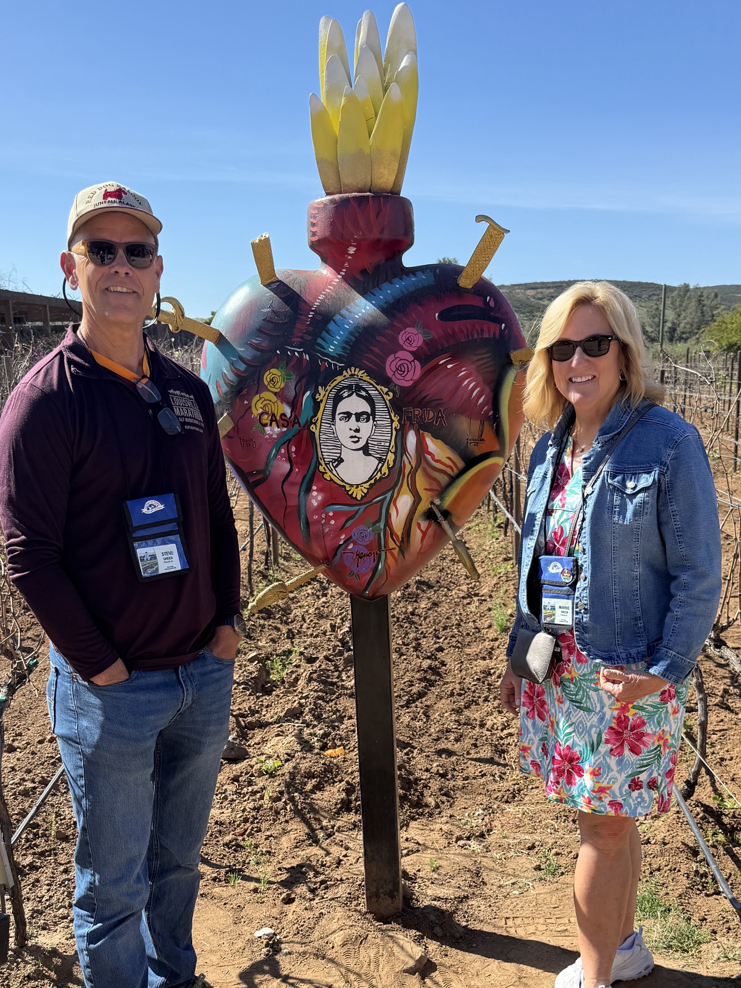 A man and a woman posing in a vineyard next to a large, colorful heart sculpture featuring a portrait, surrounded by vines and blue sky.