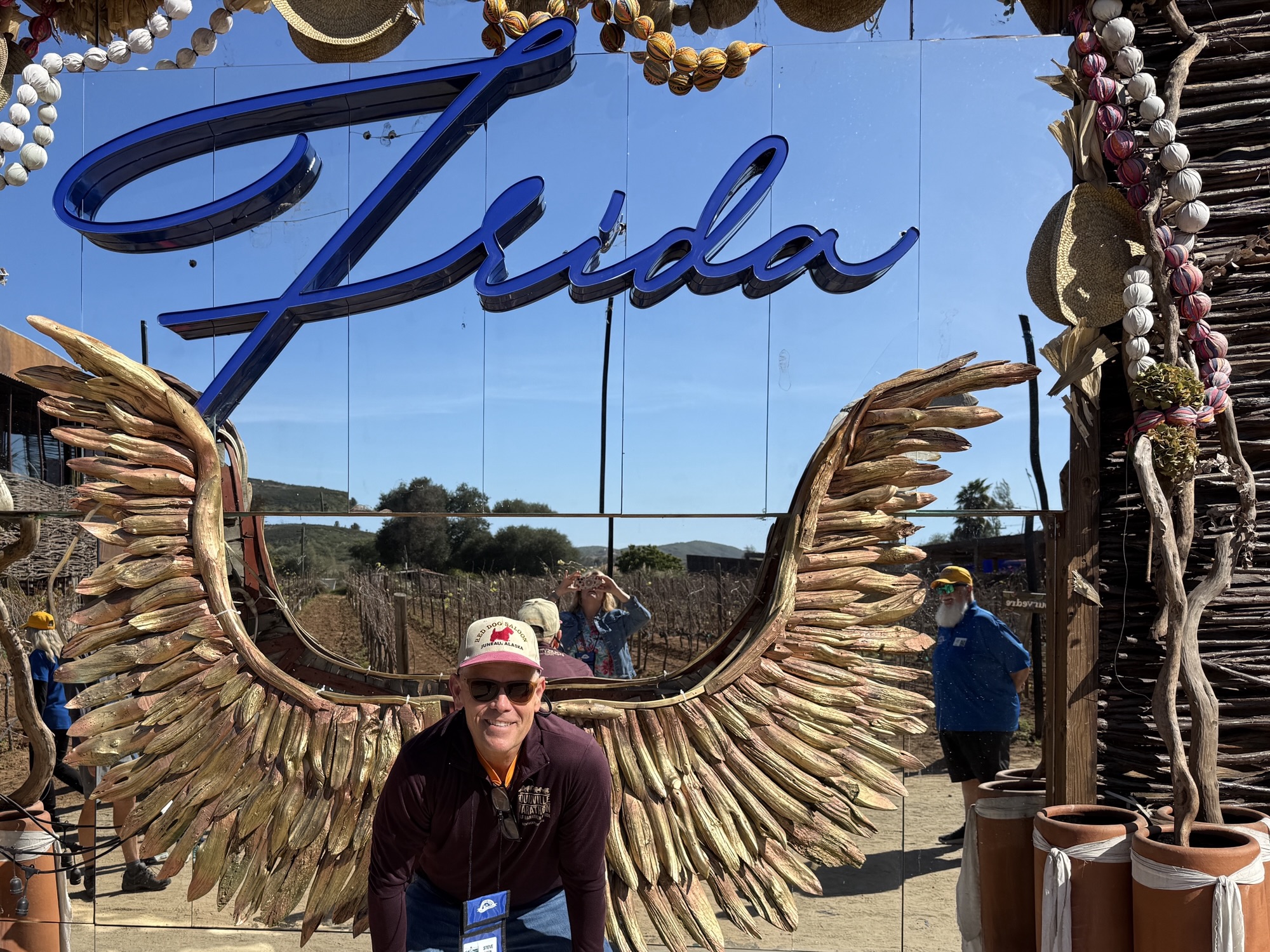 A person posing in front of a large mirror featuring the name 'Frida' and decorative wings made of leaves, with a vineyard and clear blue sky in the background.