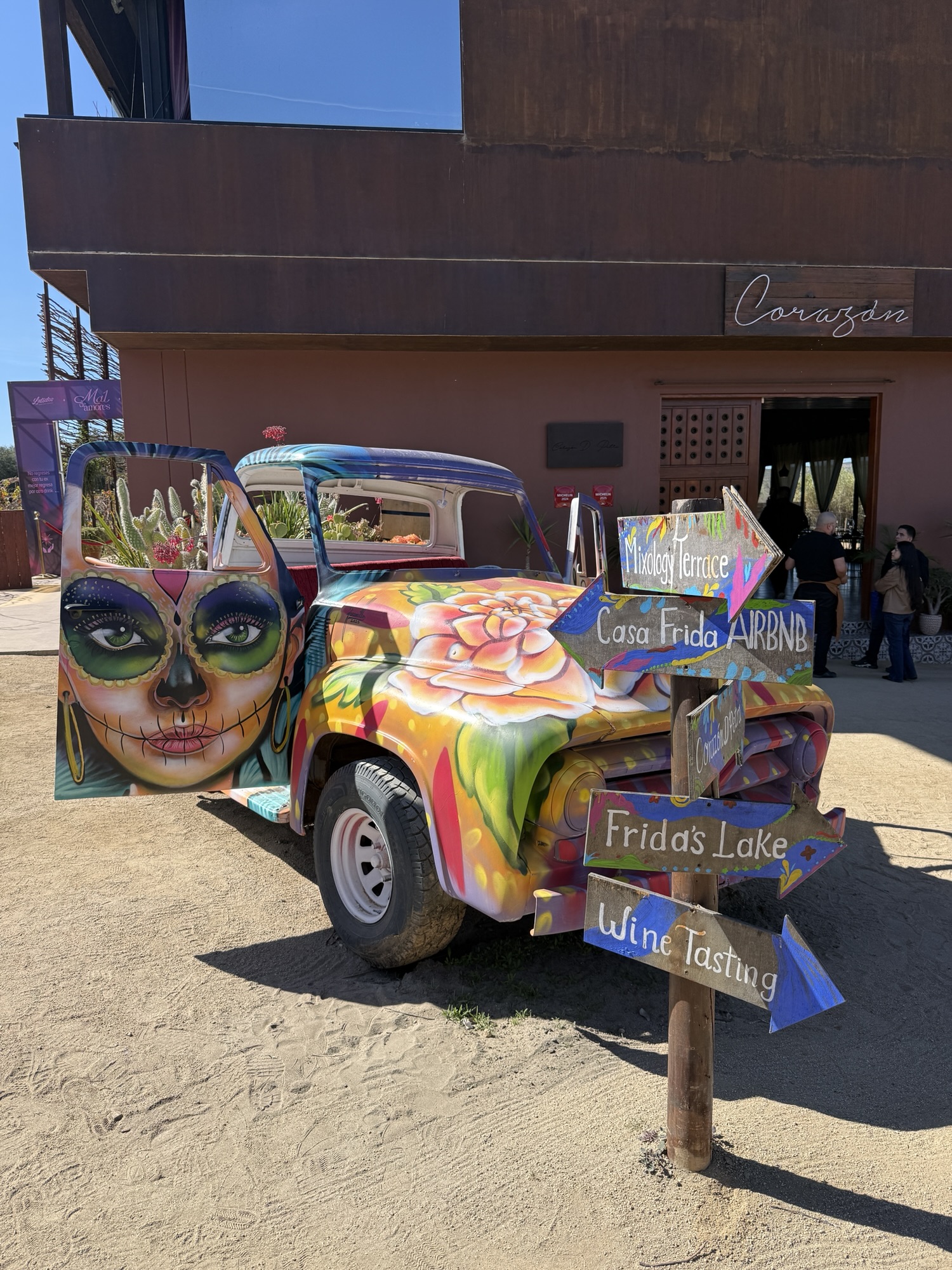 Colorfully painted vintage truck with an artistic skull design, parked in front of a building, featuring directional signs for tourist attractions like 'Frida's Lake' and 'Wine Tasting'.
