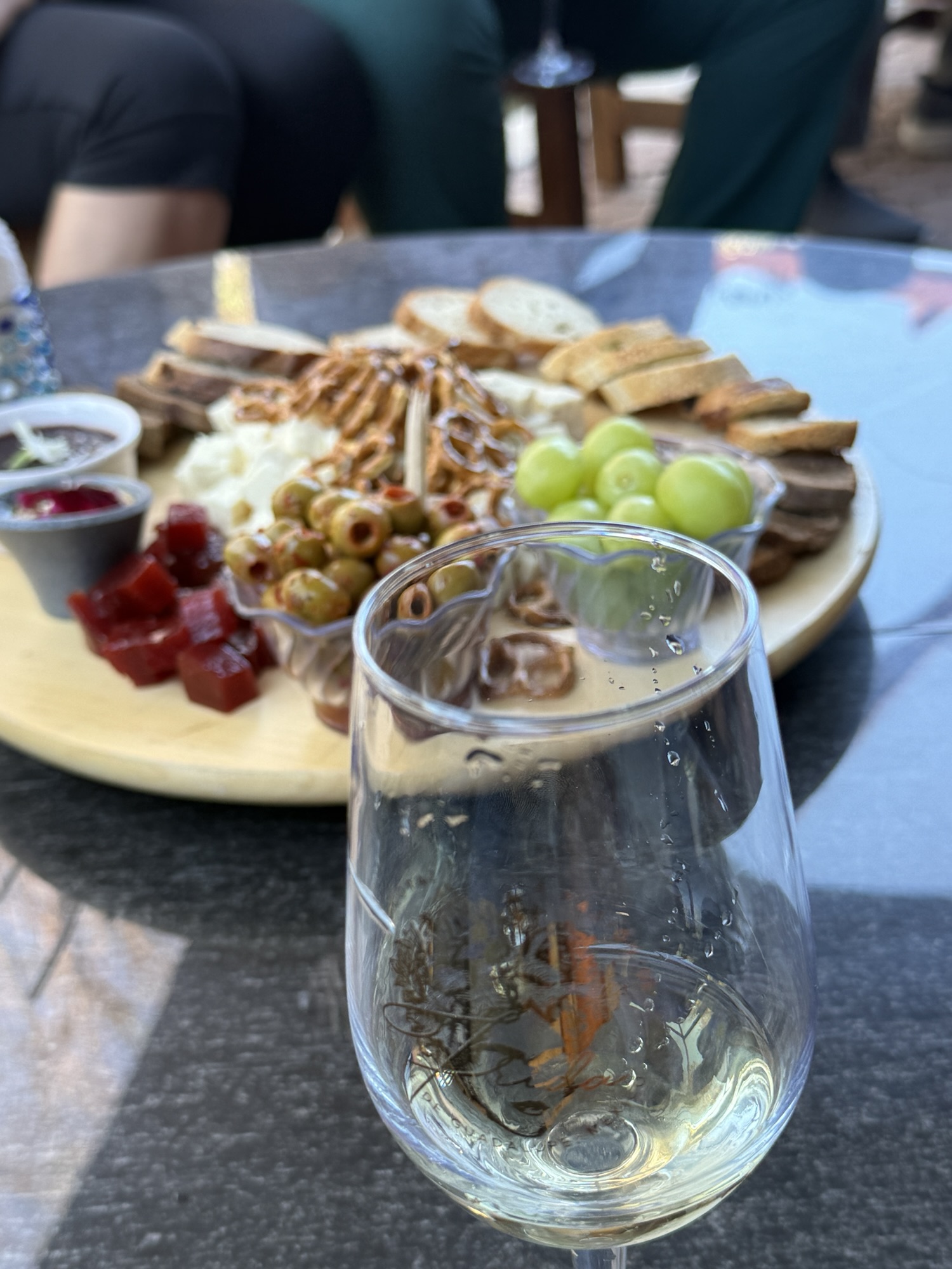 A glass of sparkling wine in the foreground, with a charcuterie board in the background featuring various cheeses, breads, olives, pretzels, and small bowls of jelly and fruit.