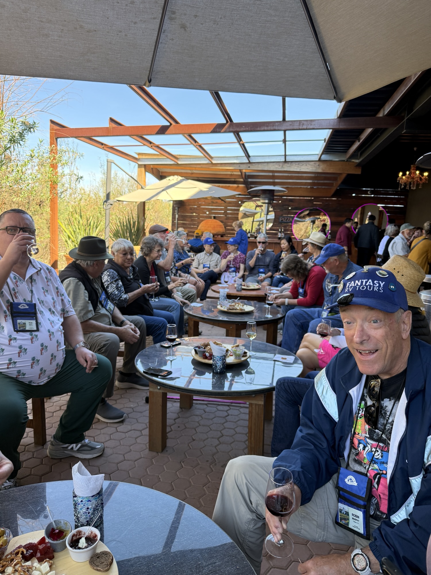 A group of people mingling on an outdoor patio during a social event, sitting around glass tables with drinks and snacks, under a shaded area.