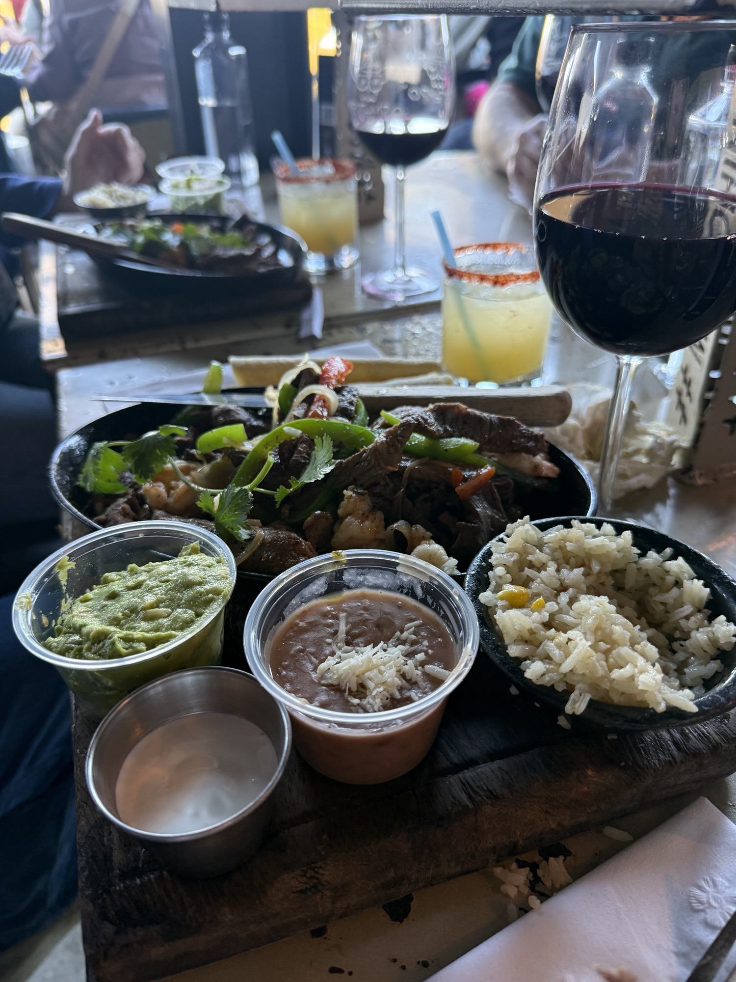 A plate of sizzling fajitas with grilled meat, bell peppers, and onions, accompanied by guacamole, refried beans, and rice, with a glass of red wine and cocktails in the background.