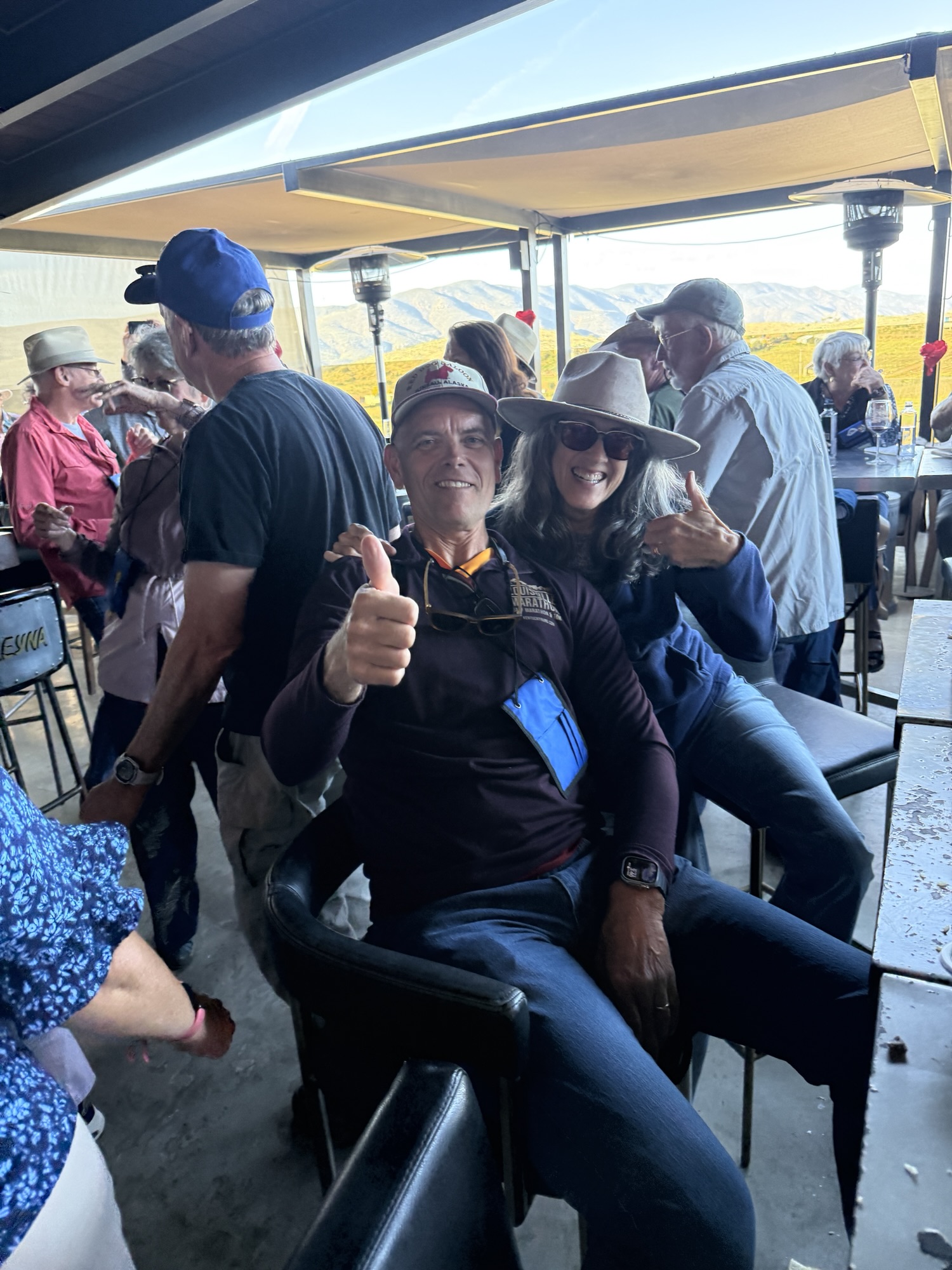 A man giving a thumbs up and a woman posing with a thumbs up, both seated at a bar with a mountain view in the background, surrounded by other people enjoying their time.