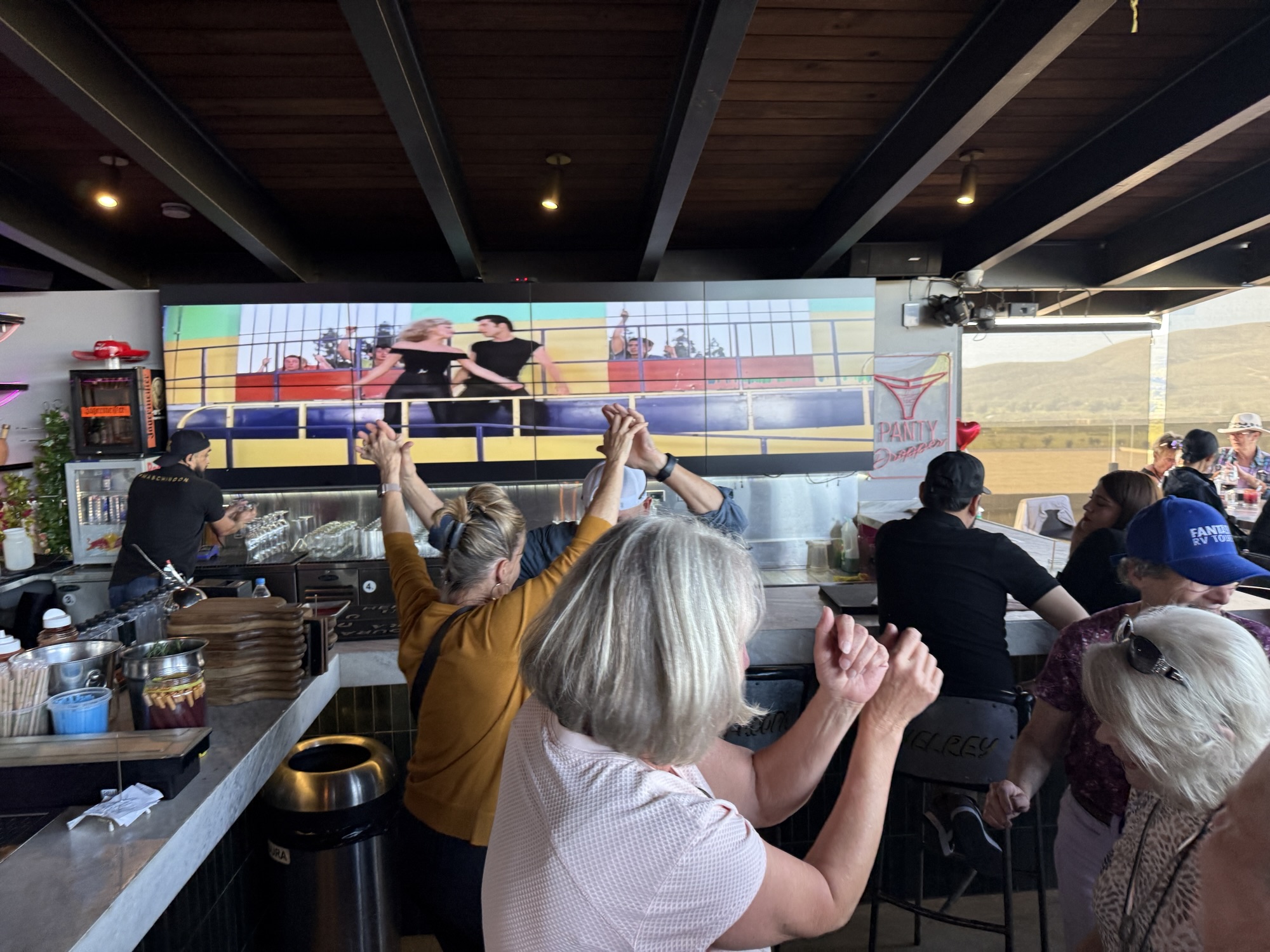A lively bar scene with patrons enjoying themselves, some dancing, and a large screen displaying a performance in the background.