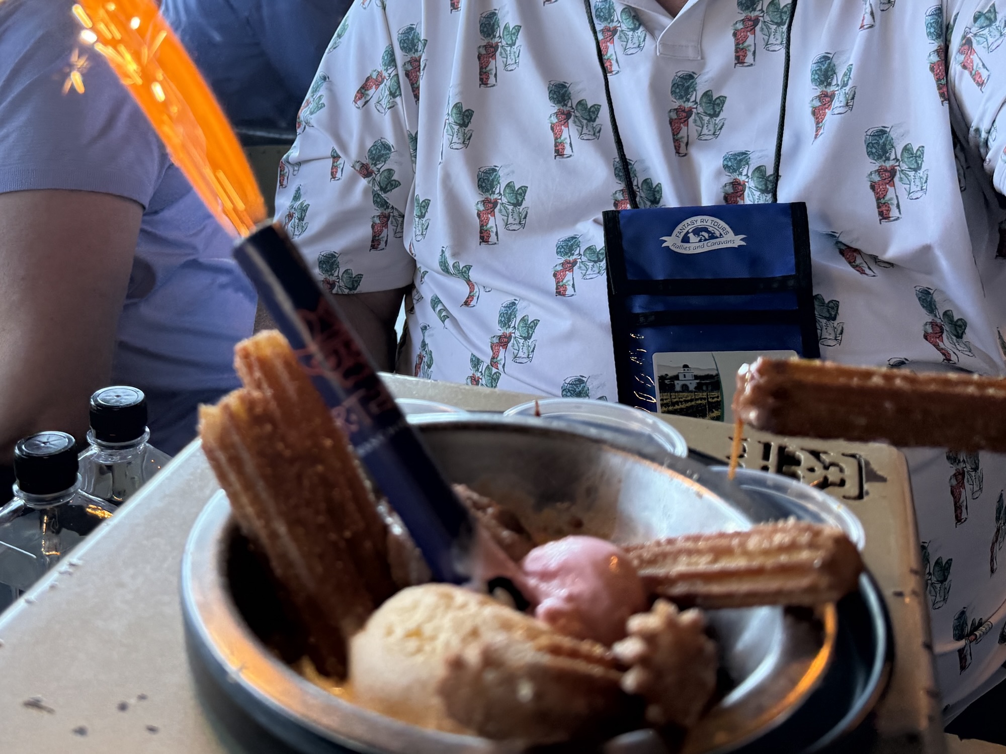 A dessert dish featuring churros and ice cream, topped with a sparkler, served on a table with a person in a colorful shirt in the background.