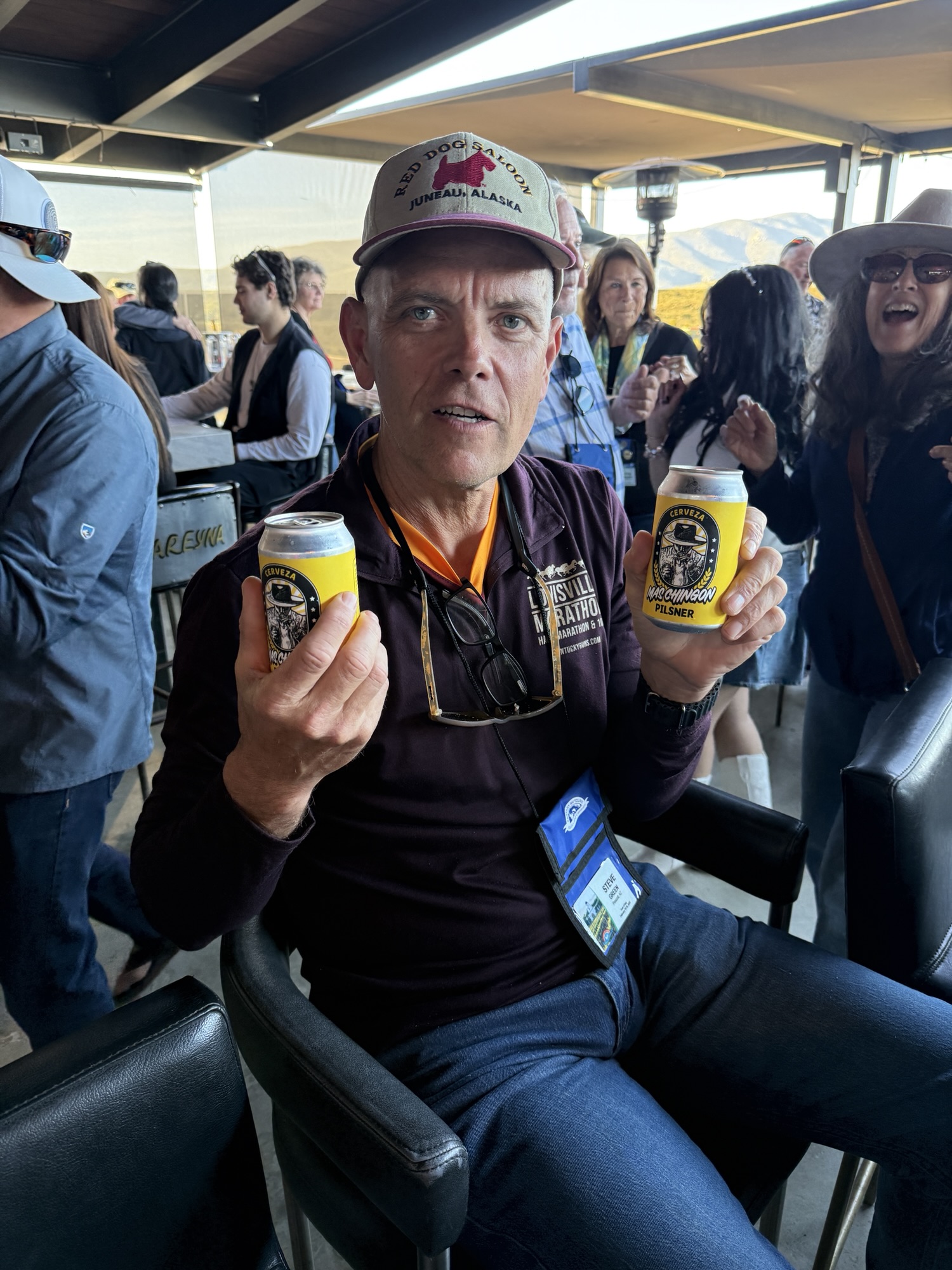A man sitting in a bar holding two cans of beer, wearing a hat with 'Red Dog Saloon Juneau Alaska,' and a lanyard with a name tag. Other people are seen in the background, enjoying a social gathering.
