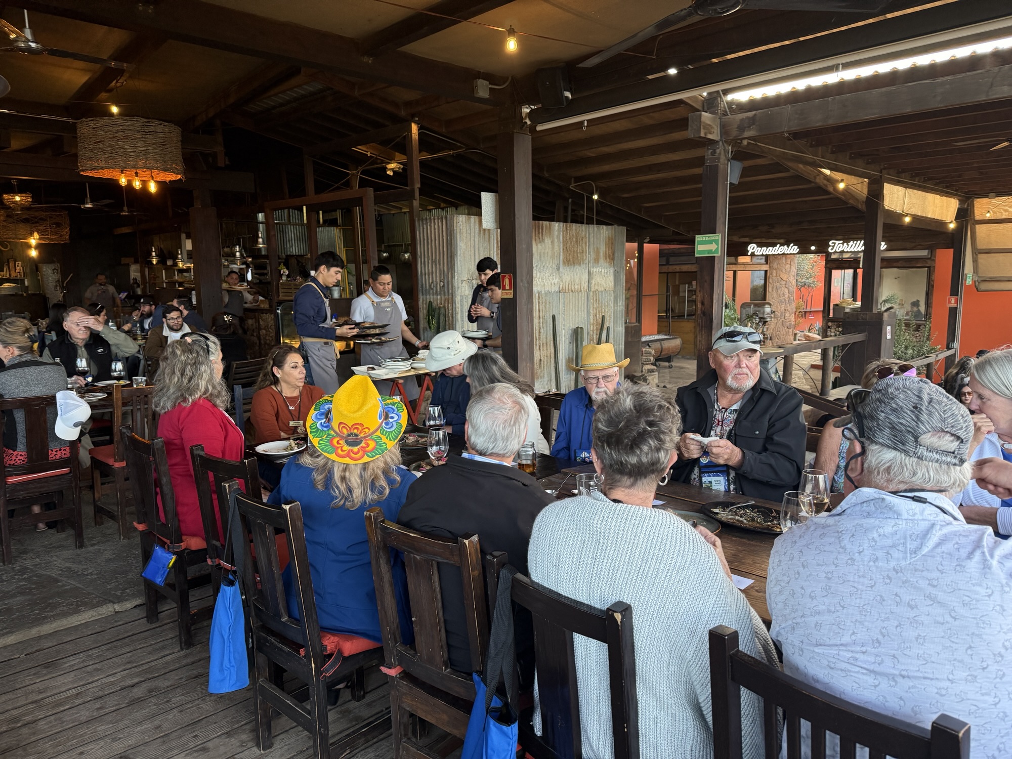 A lively restaurant scene with a group of diverse diners gathered around a table, enjoying their meals, while staff serve food in a rustic setting.