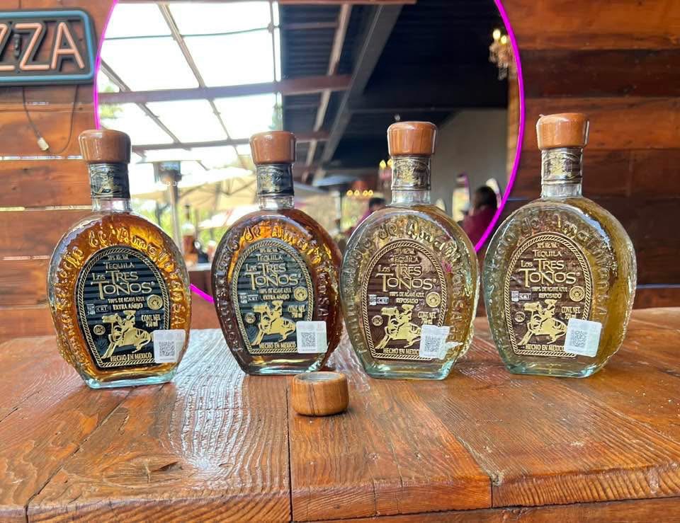 Four bottles of Tequila 'Los Tres Tonos' in various types (Extra Añejo, Añejo, Reposado) displayed on a wooden table, with a blurred restaurant background.