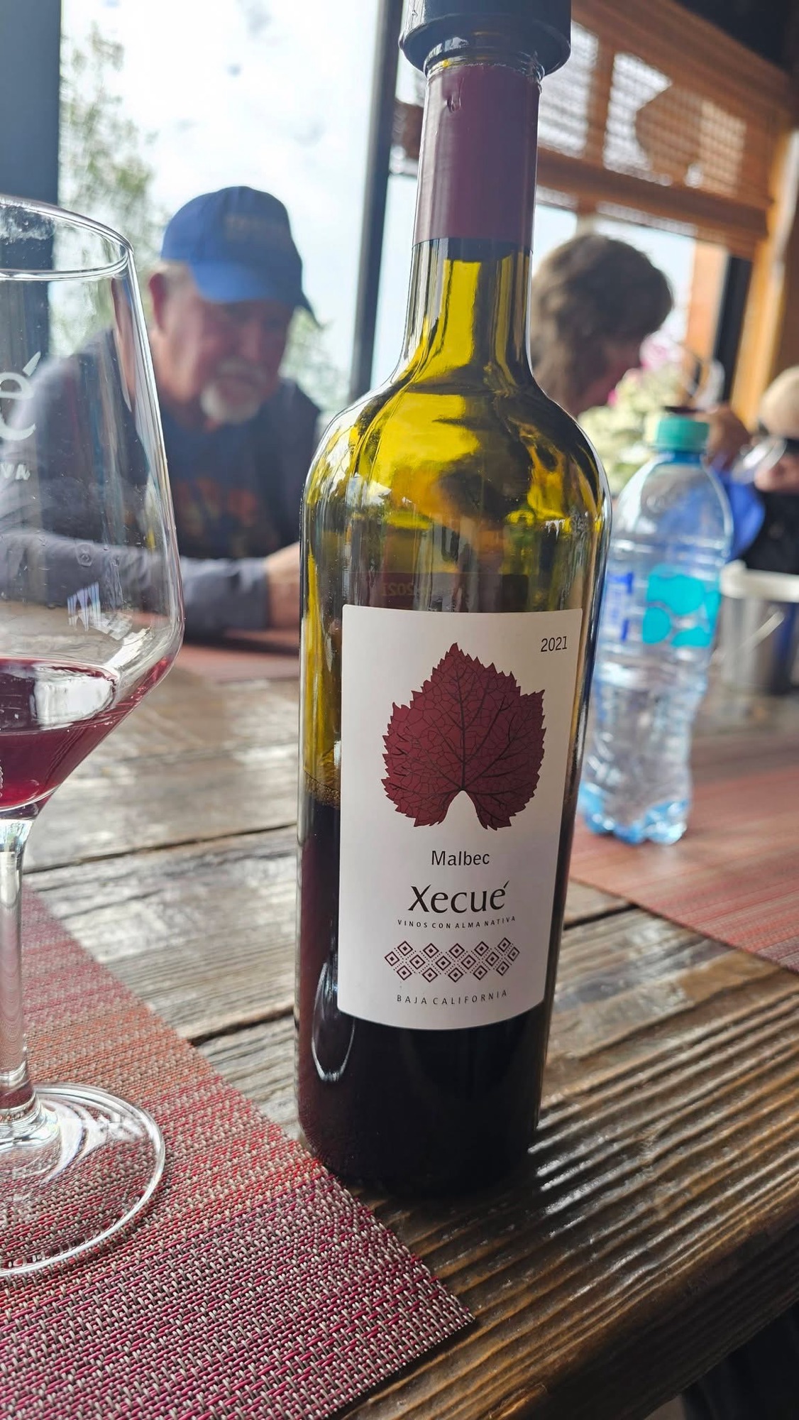 A bottle of Xecué Malbec wine from Baja California, 2021 vintage, displayed on a wooden table with a wine glass partially filled with red wine. A blurred background shows a person wearing a blue hat.