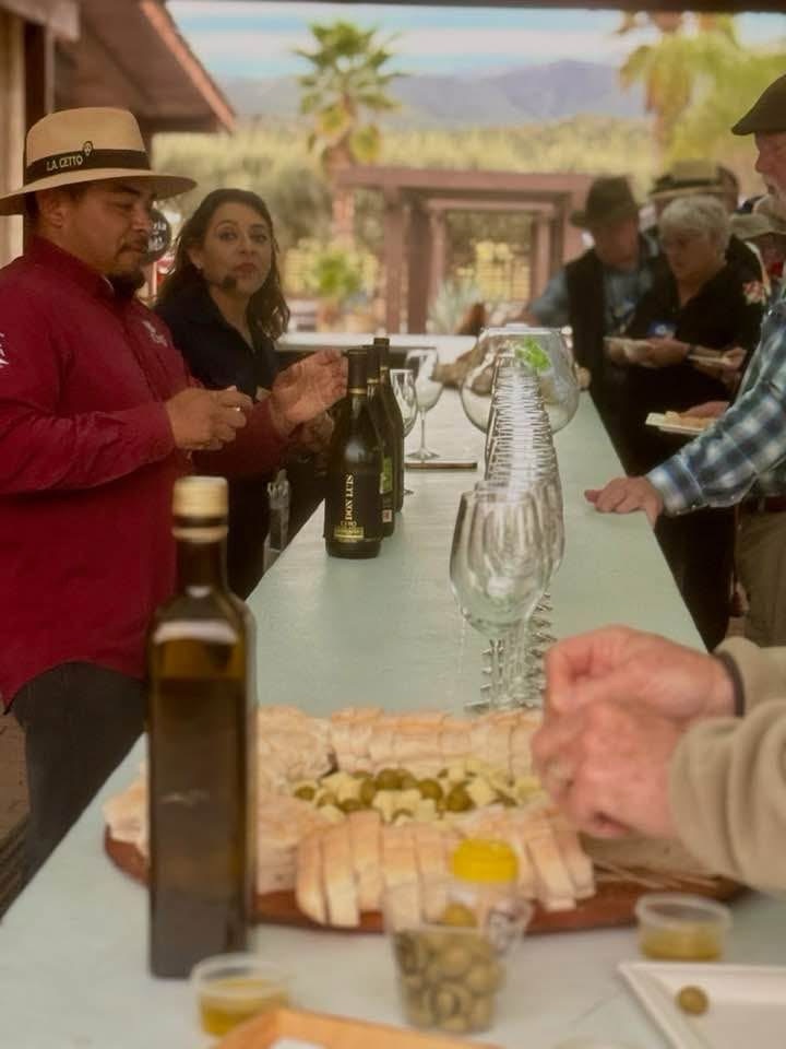 A group of people enjoying a tasting event at a long table with various food items, including sliced bread and olives, while an instructor explains the offerings. Bottles of wine are displayed on the table.