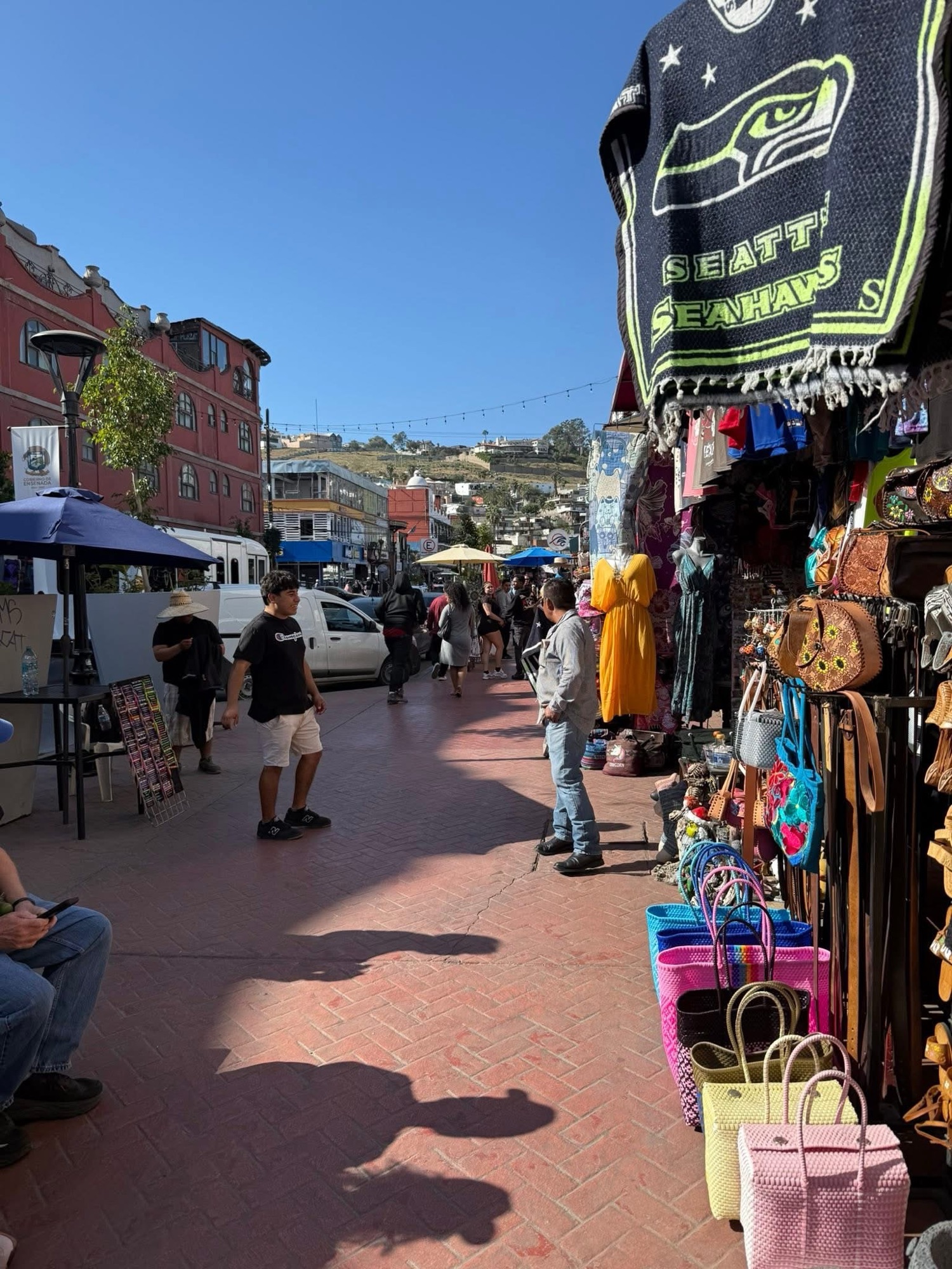 A bustling street market scene with colorful stalls displaying various goods, including handbags and clothing. People are walking and browsing, with a clear blue sky overhead.