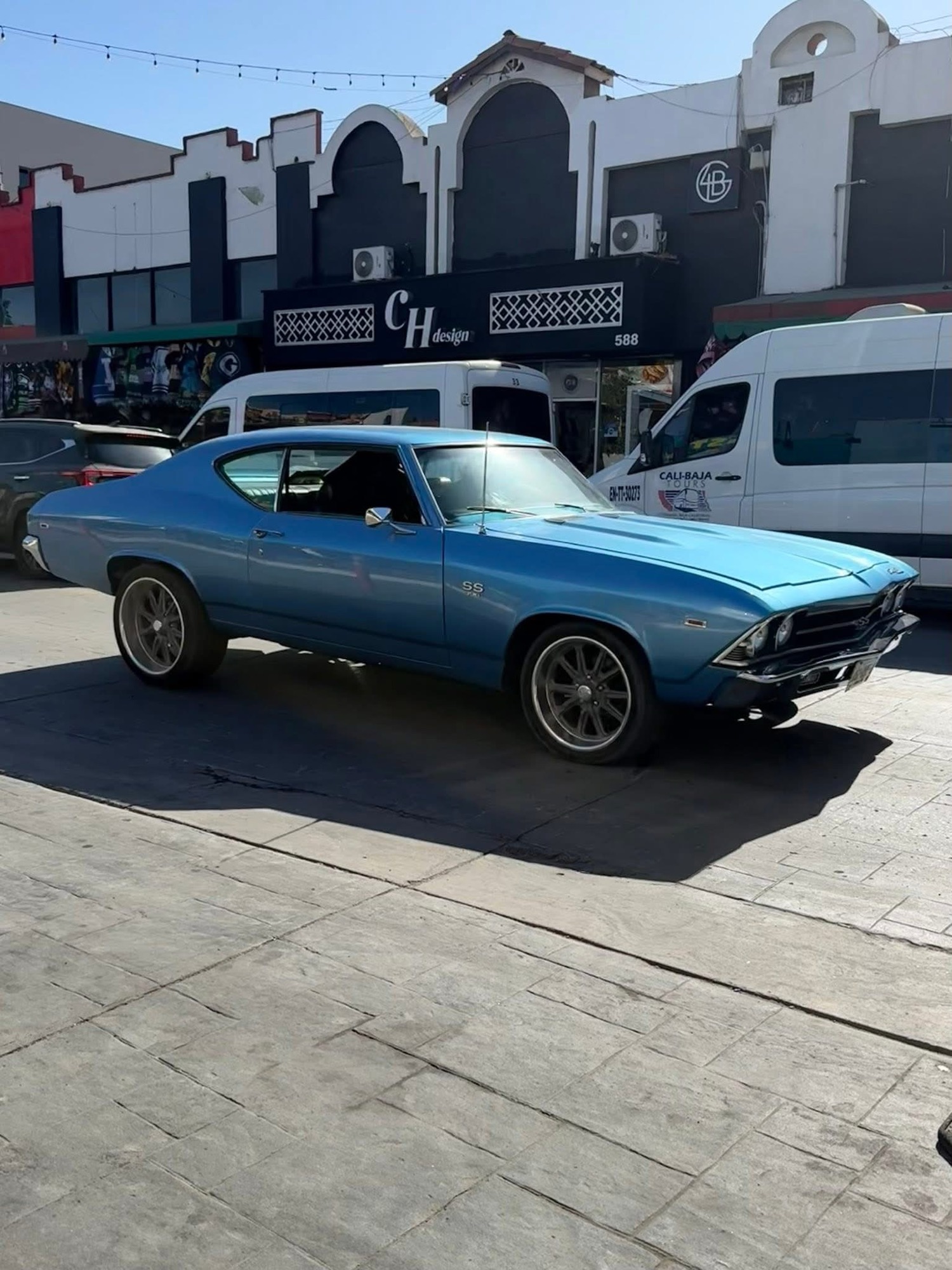 A classic blue Chevrolet Chevelle SS parked on a busy street with shops in the background.