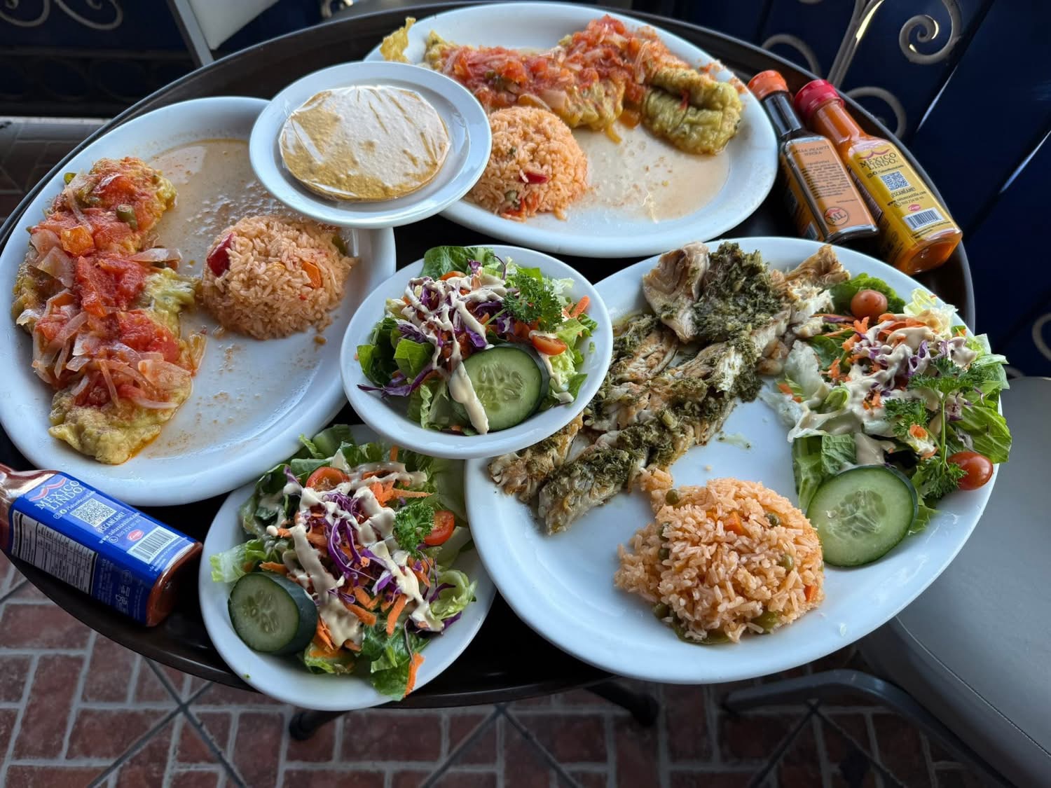 A table set with plates of colorful Mexican food, including enchiladas topped with sauce, rice, grilled fish, and fresh salads, accompanied by various hot sauces and beverages.