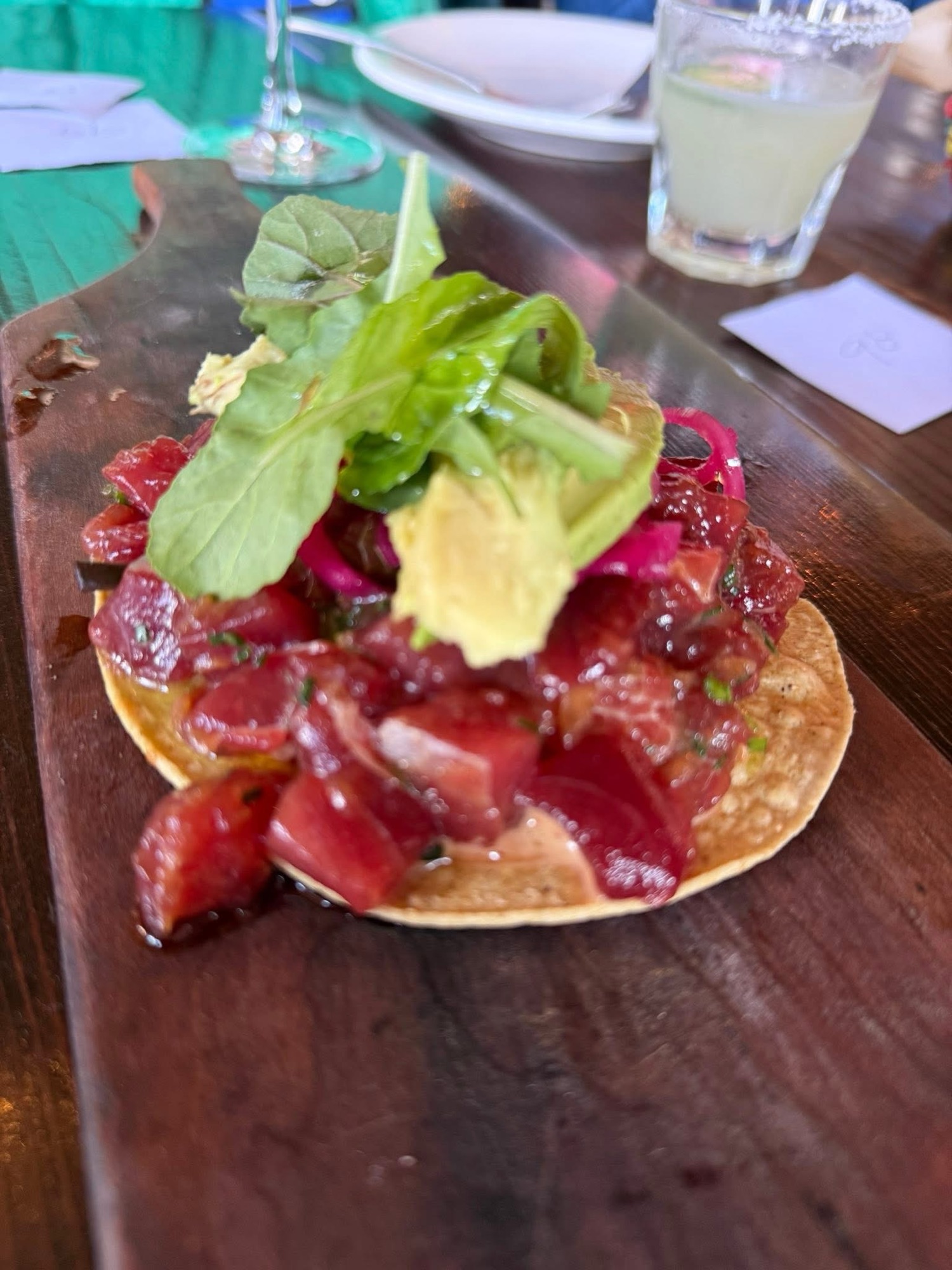 A wooden platter displaying a vibrant taco topped with fresh diced fish, avocado slices, pickled onions, and leafy greens.
