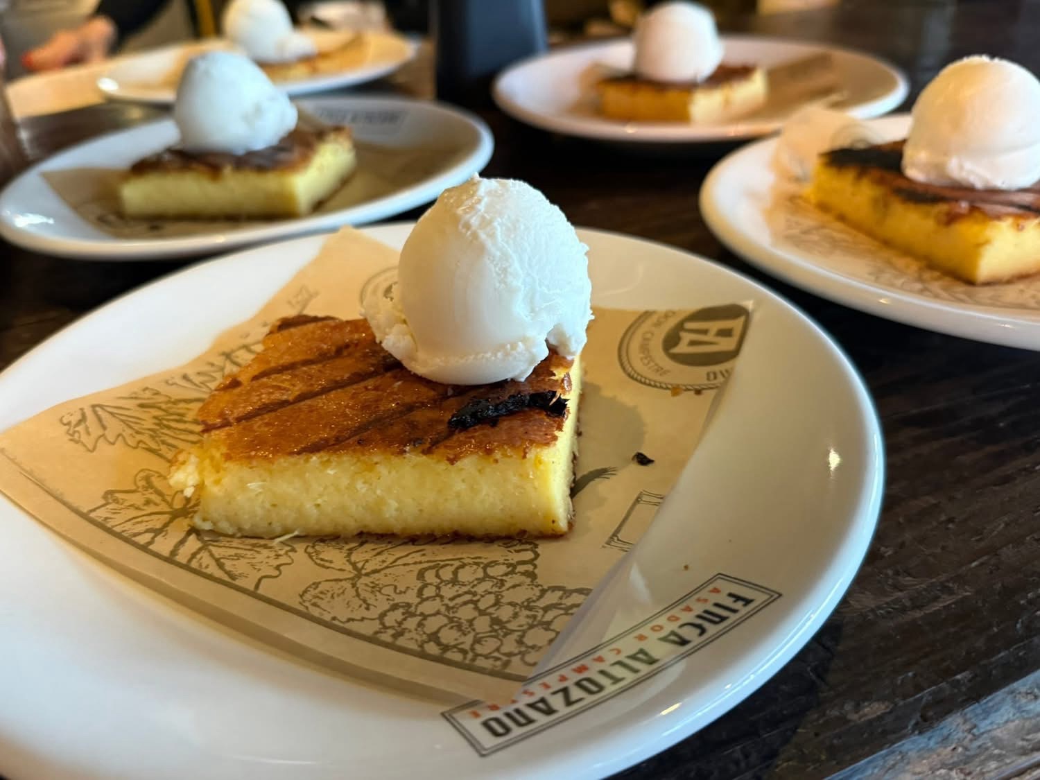 A slice of golden dessert topped with a scoop of vanilla ice cream, served on a decorative plate with a paper liner.