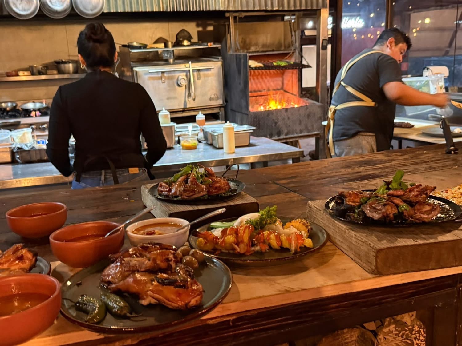 A busy restaurant kitchen scene featuring two chefs at work, with a variety of grilled meats and colorful dishes served on wooden tables in the foreground.