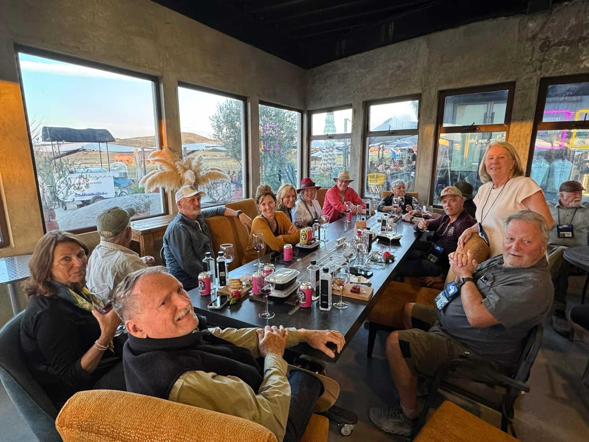A group of people gathered around a long table in a cozy indoor setting, enjoying drinks and conversation. The room features large windows showcasing an outdoor view, with guests smiling and engaging with one another.