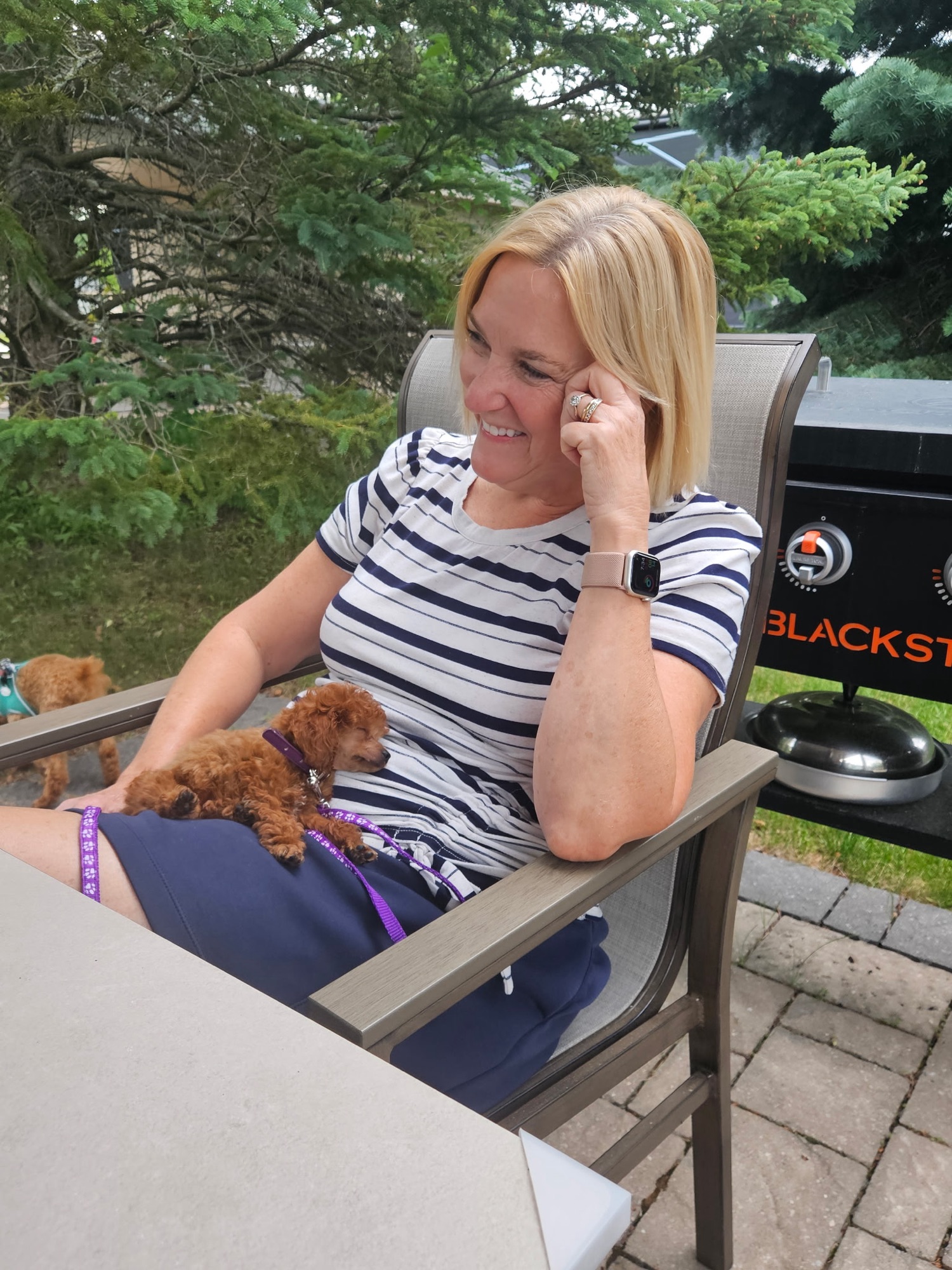 A woman sitting on a chair outdoors, smiling and resting her chin on her hand, with a small sleeping dog on her lap. The background features green trees and an outdoor grill.