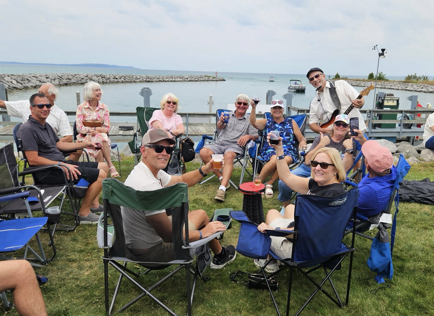 A group of people enjoying a gathering outdoors by the water, sitting in folding chairs, toasting with drinks, and smiling. A musician is playing a guitar in the background.