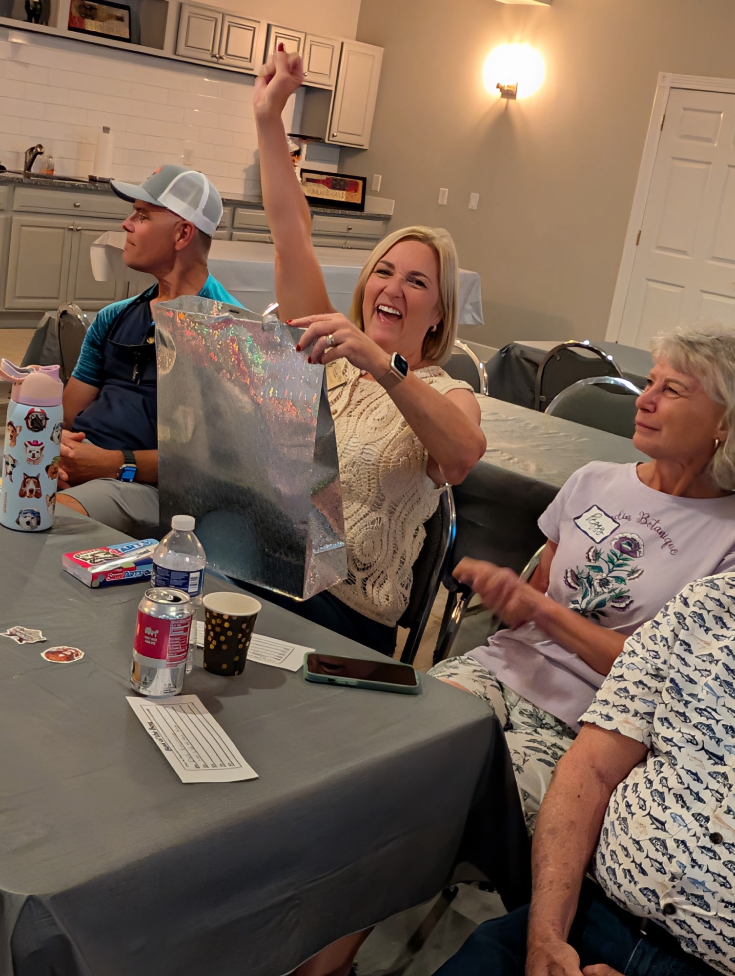 A woman joyfully raises her arm while opening a gift at a gathering, surrounded by other attendees at a table with snacks and drinks.