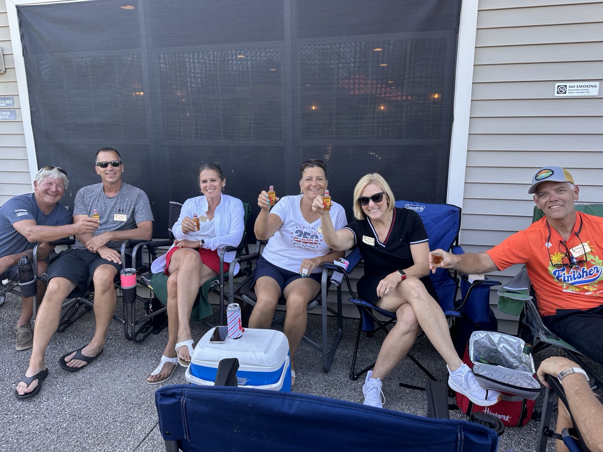 A group of five people sitting in camping chairs, smiling and holding drinks outdoors, with a cooler nearby.