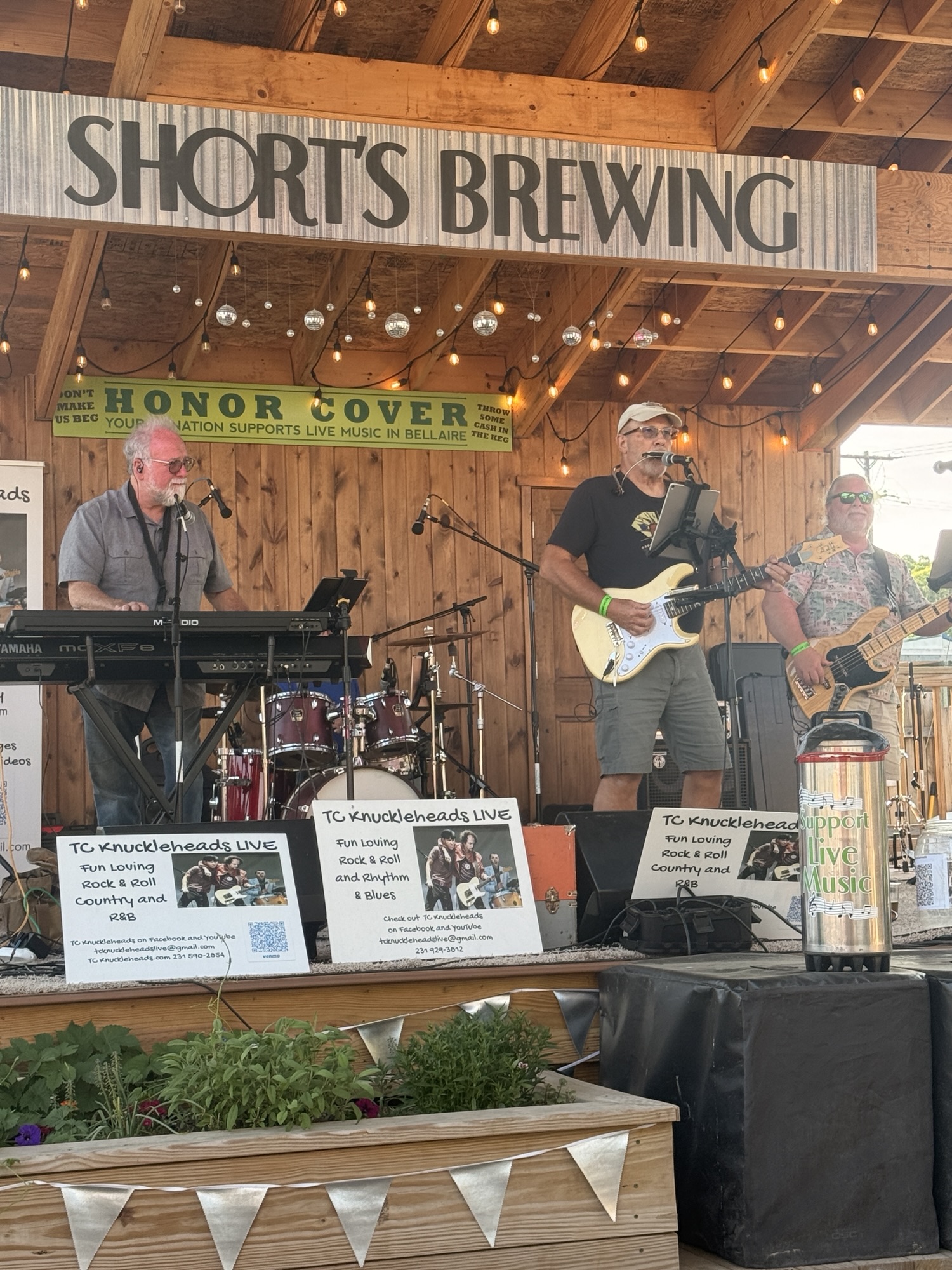 A live band performing at Shorts Brewing in Bellaire, featuring musicians with instruments, vibrant decorations, and promotional signs for TC Knuckleheads.