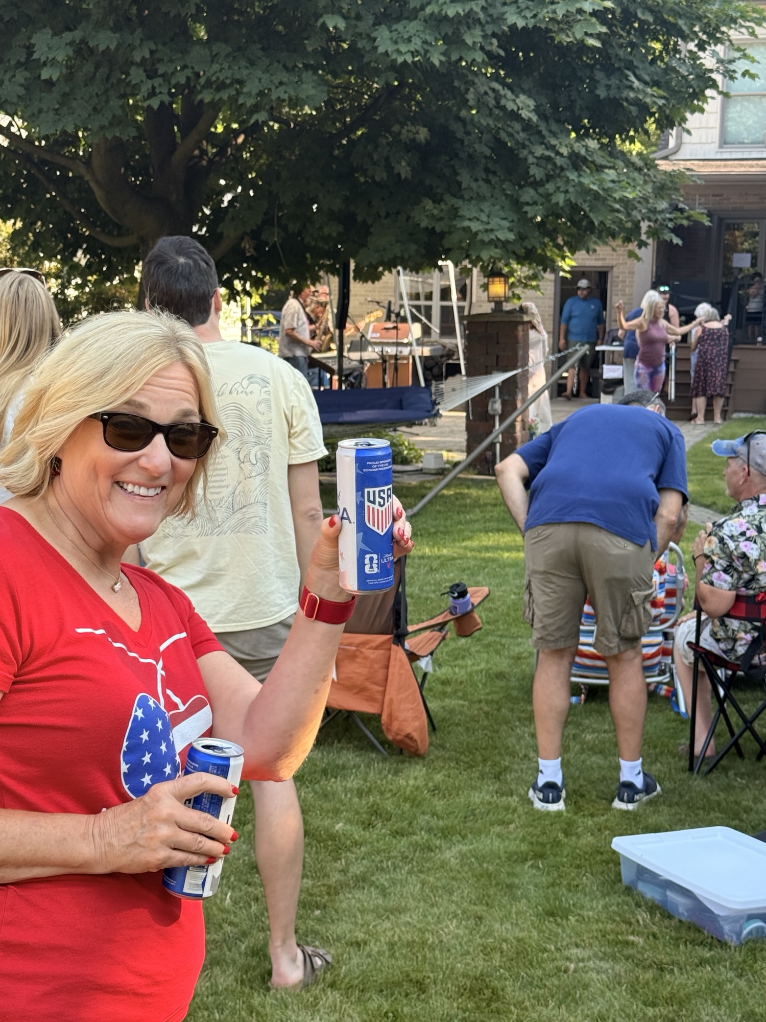 A woman in a red shirt with a heart design is smiling and holding two cans of beer at an outdoor gathering, with people socializing in the background.