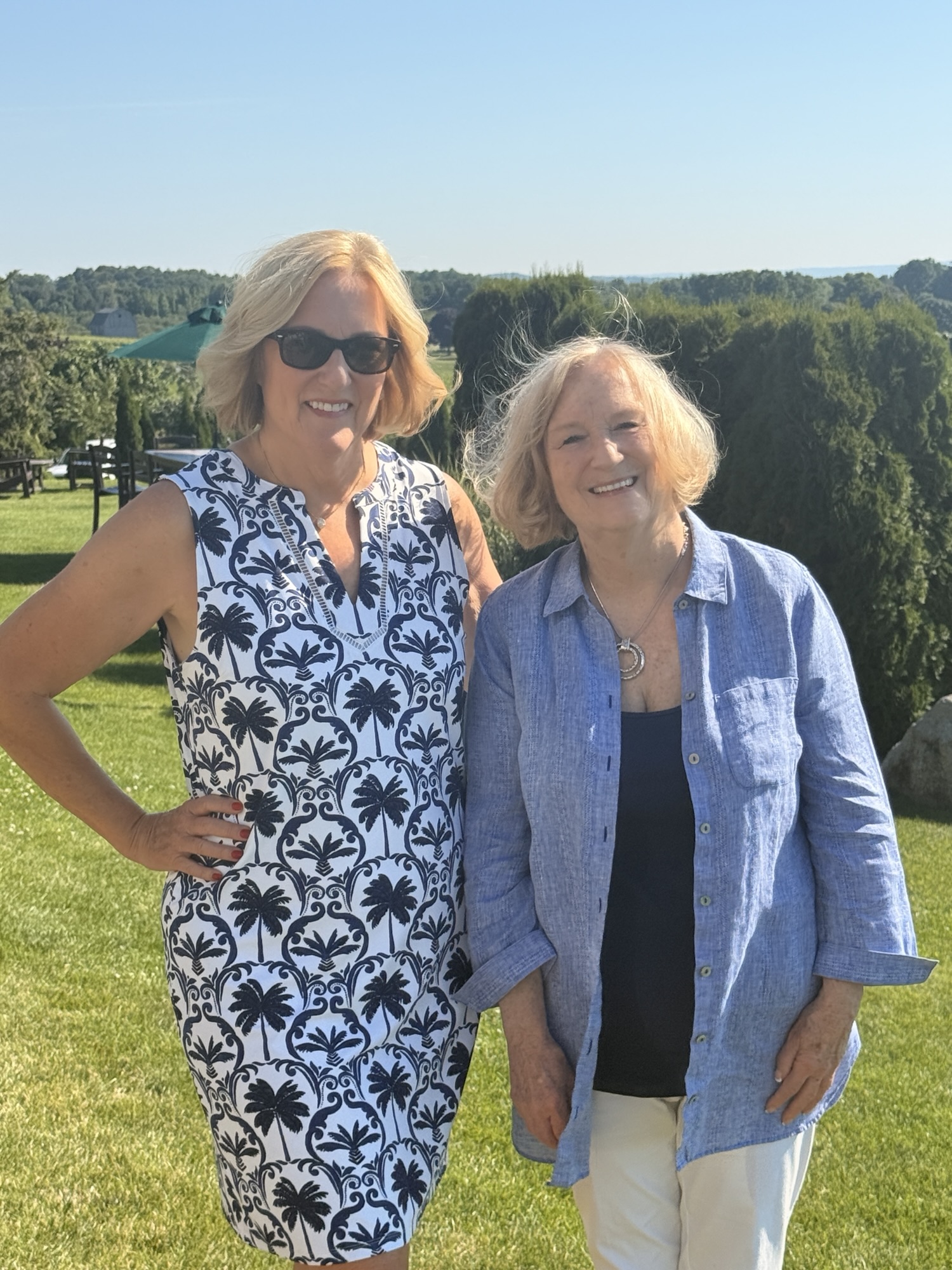 Two women smiling together outdoors on a sunny day, with green grass and trees in the background.