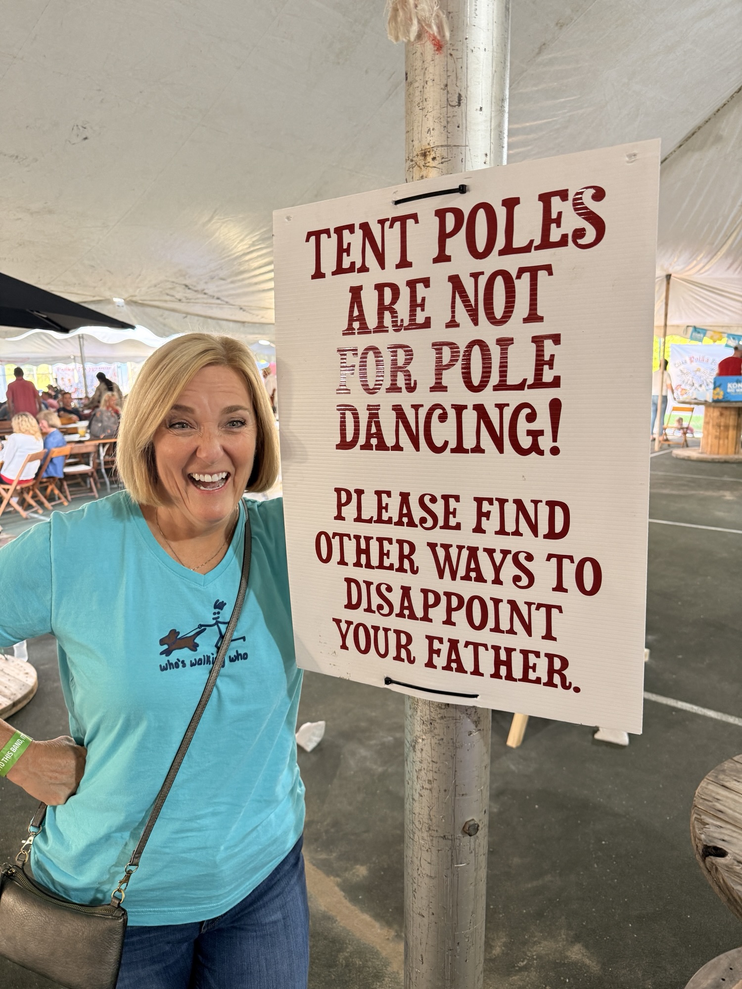 A woman smiles and poses beside a humorous sign that reads 'TENT POLES ARE NOT FOR POLE DANCING! PLEASE FIND OTHER WAYS TO DISAPPOINT YOUR FATHER.'