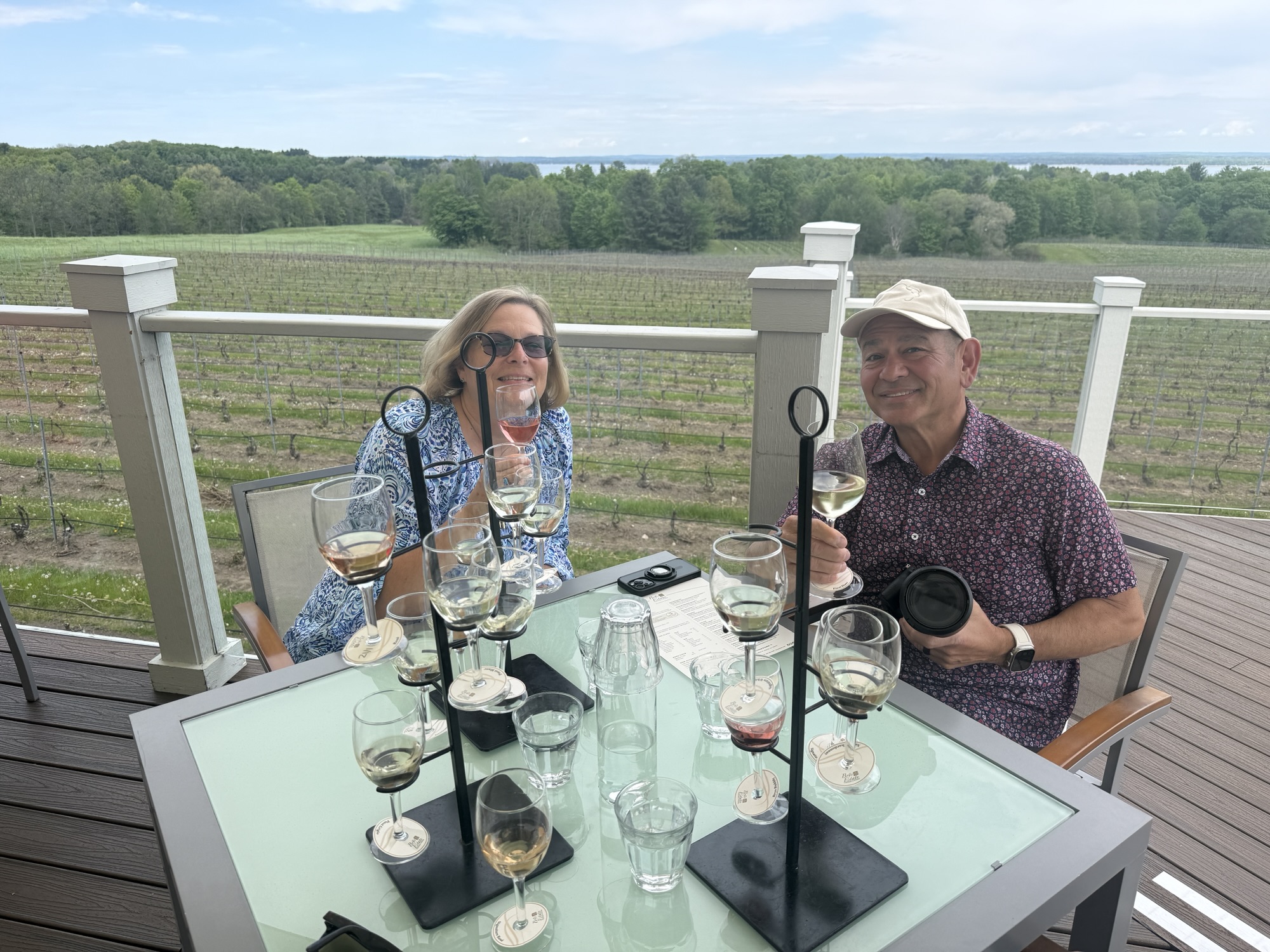A woman and a man enjoying wine tasting at a vineyard, seated at a table with various wine glasses, overlooking a lush green vineyard landscape.