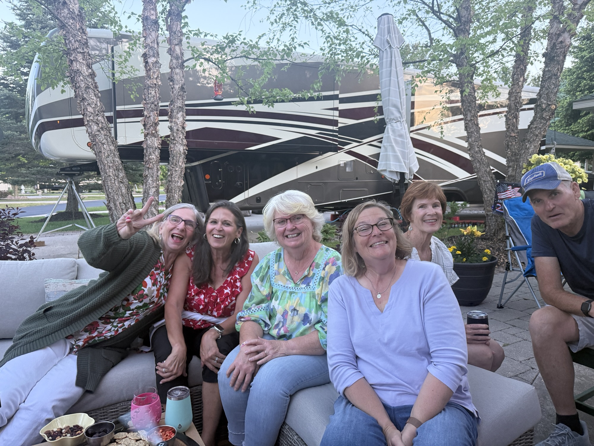 A group of six people smiling and posing together on a patio, with a large RV in the background and trees around them.