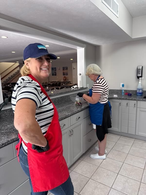 Two women working in a kitchen; one is smiling and wearing a red apron, while the other is preparing food at a counter.