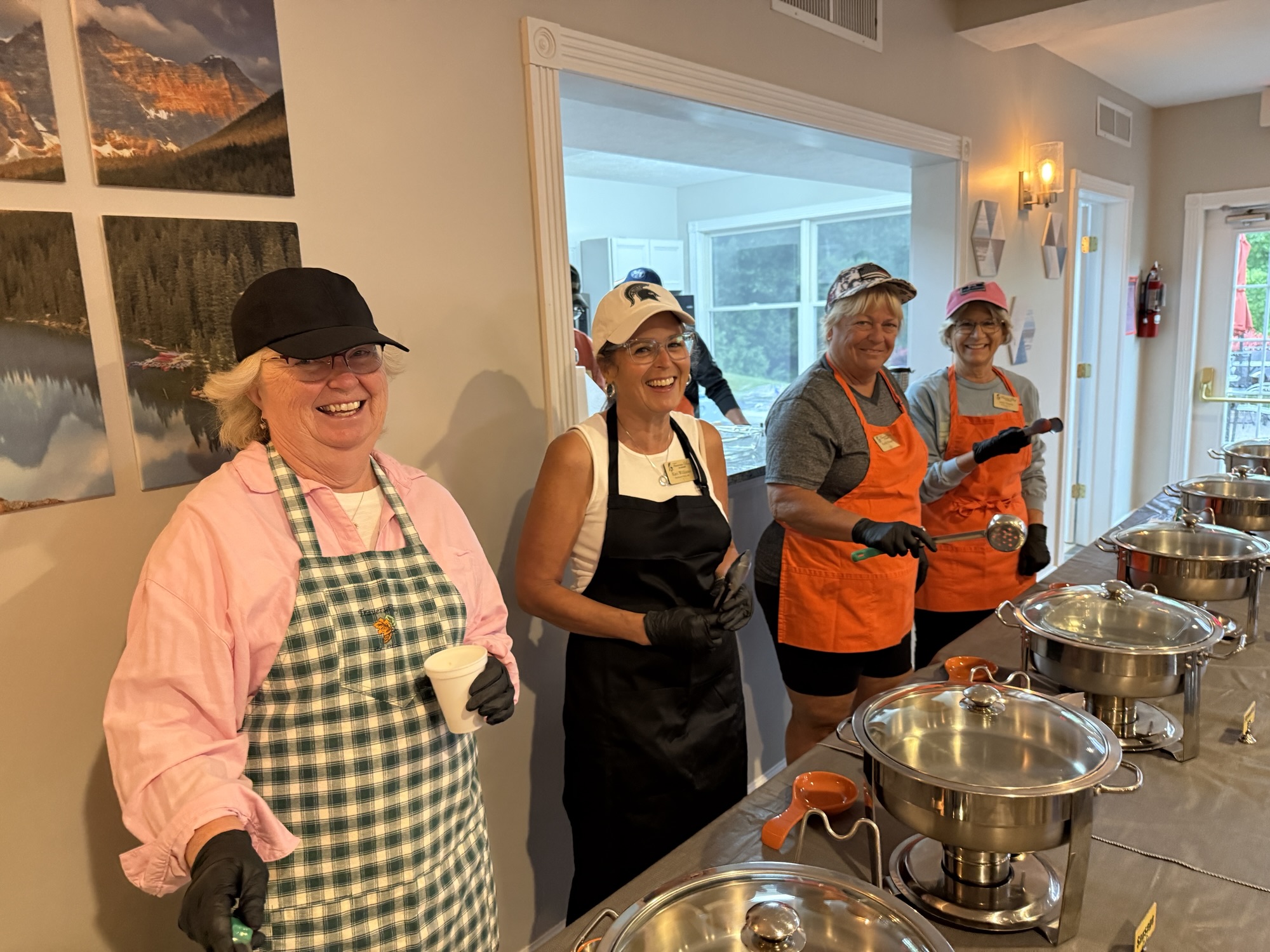 A group of four women smiling and working in a kitchen, wearing aprons and gloves, standing behind a table with several pots and cooking utensils.