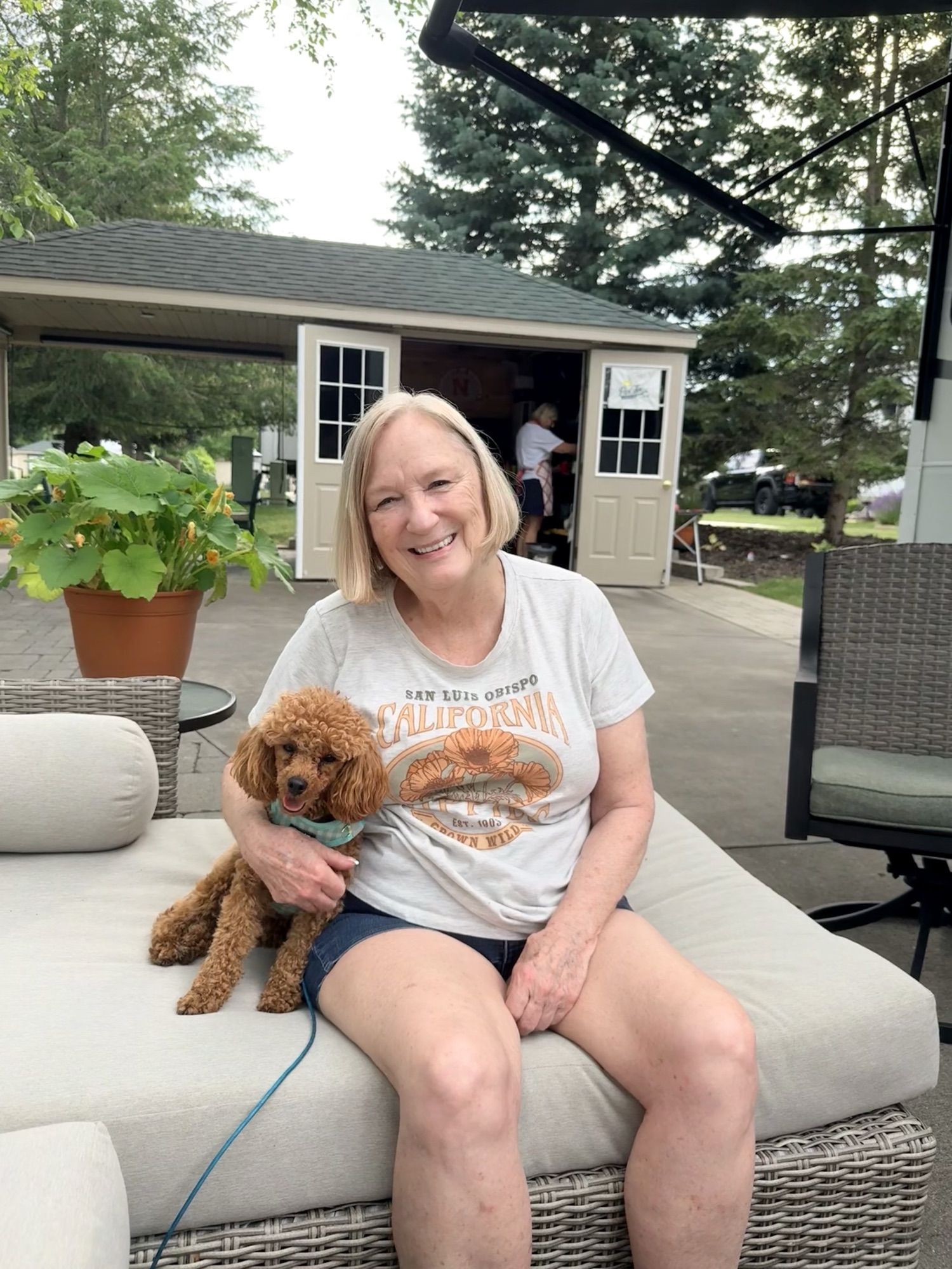 A smiling woman sitting on a sofa with a small, fluffy dog next to her, outdoors in a garden setting with trees and a shed in the background.
