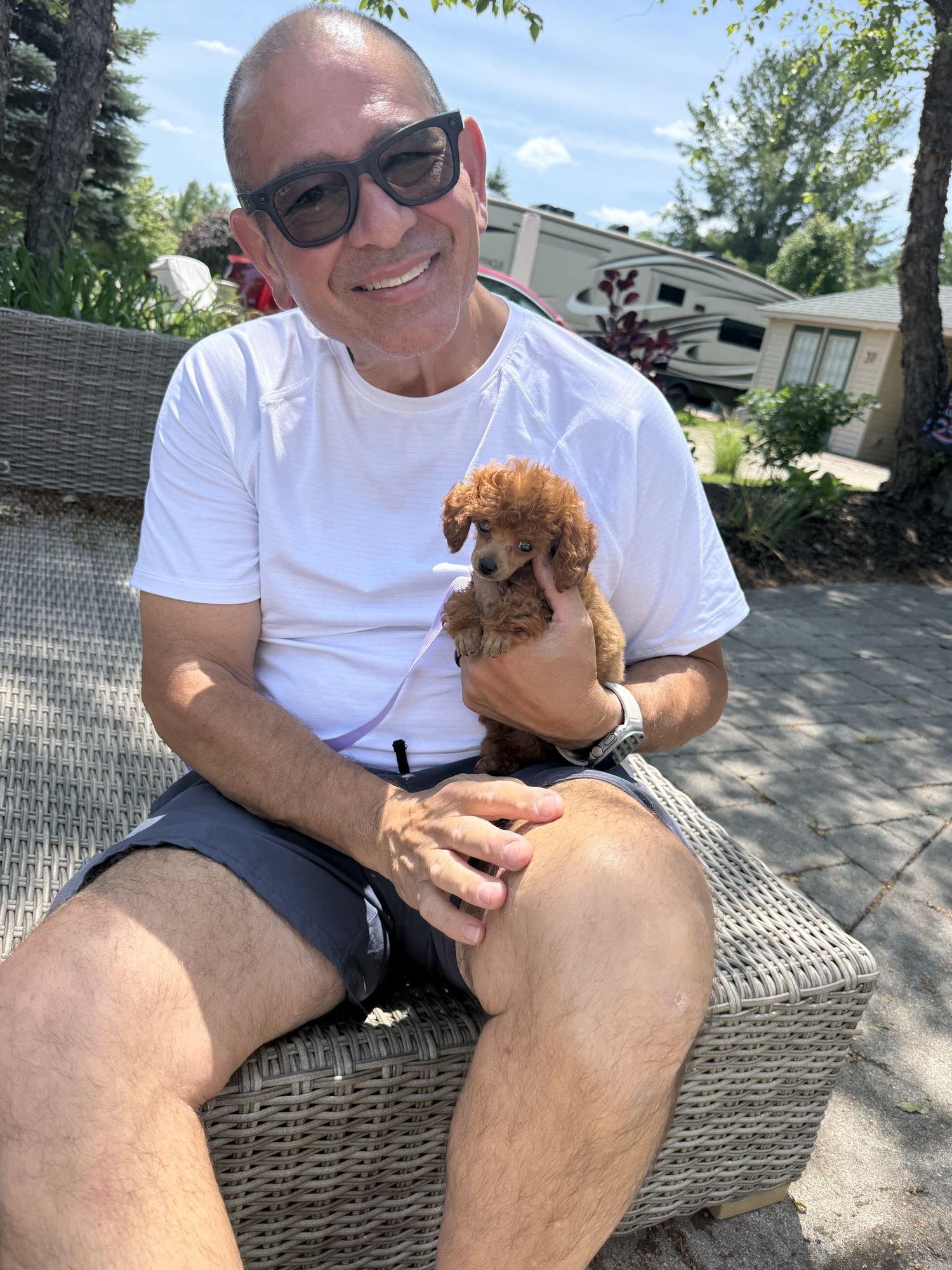 A smiling man wearing sunglasses holds a small, fluffy brown puppy in his arms while sitting on a wicker bench outdoors on a sunny day.