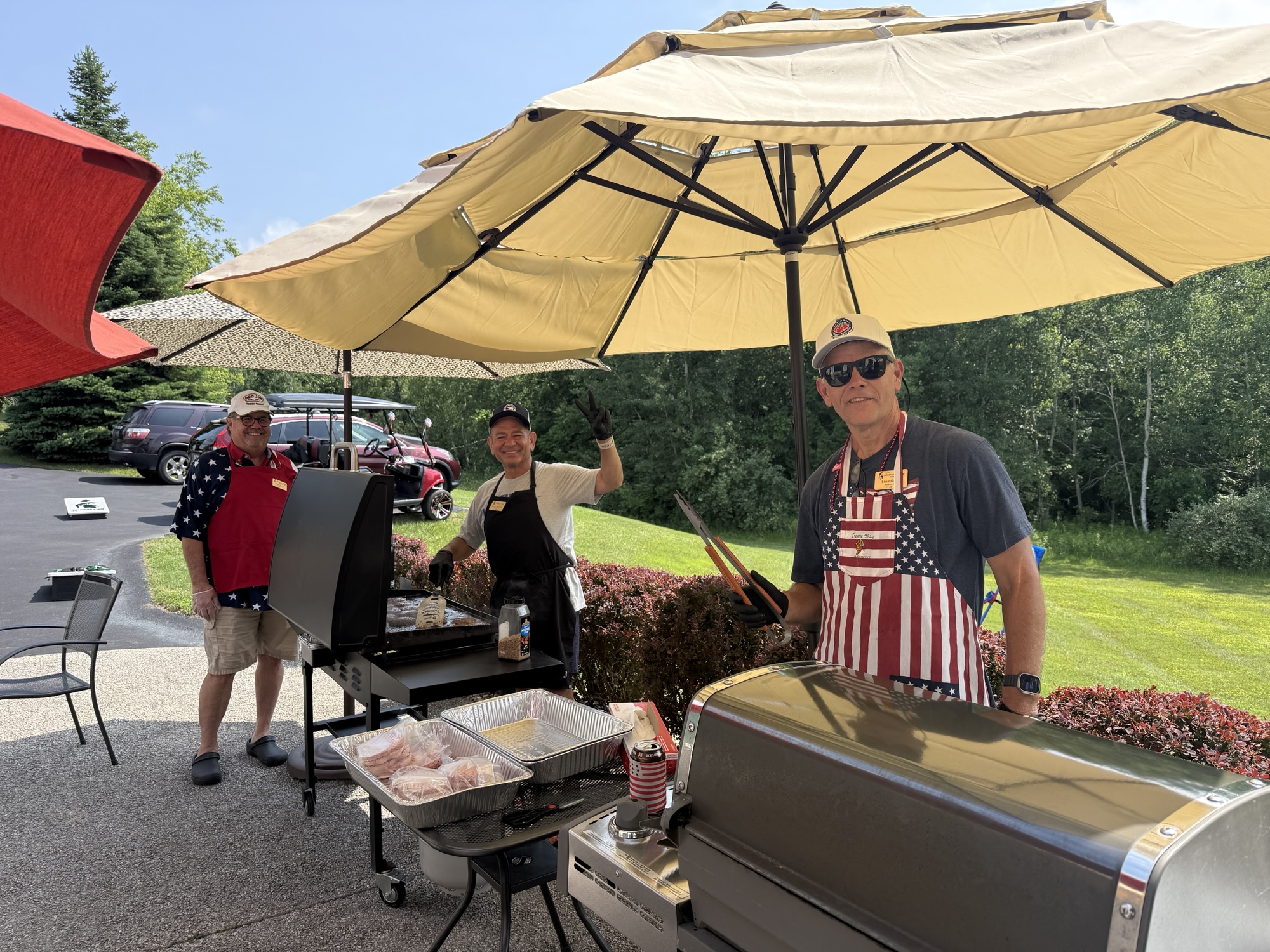 Three individuals grilling outdoors under large umbrellas, with food on the grill and a grassy area in the background.