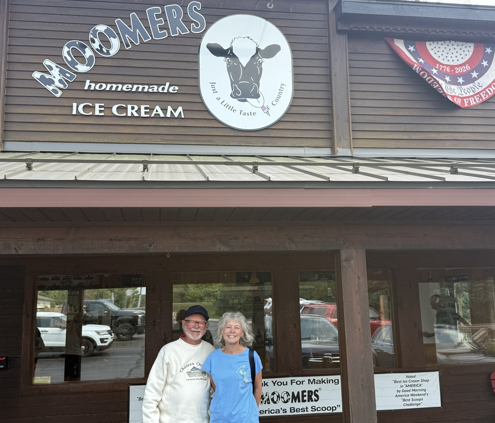 A man and woman smiling in front of Moomers homemade ice cream shop, featuring a wooden exterior and a sign with a cow logo.