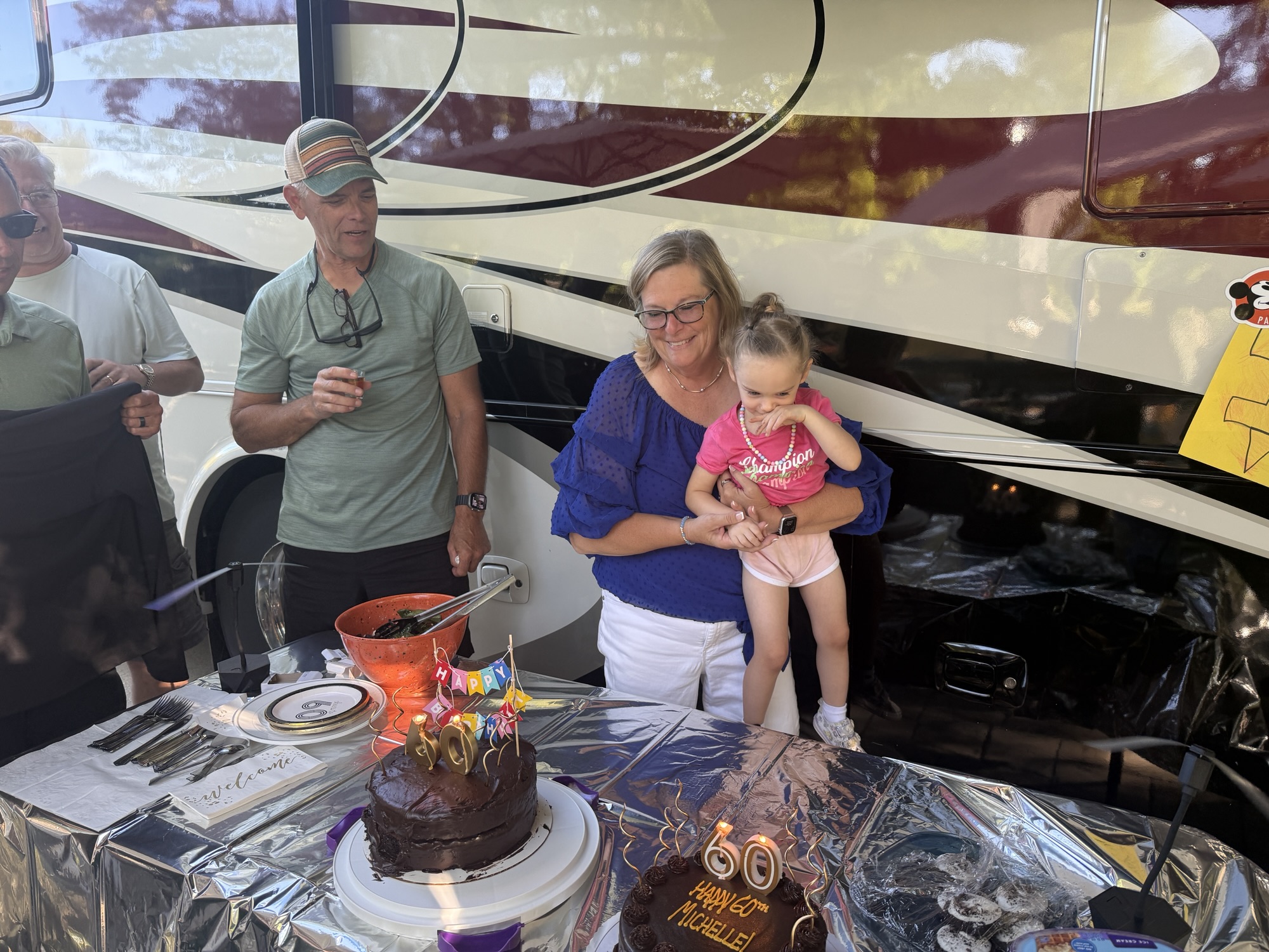 A woman holding a young girl stands next to a decorated cake for a 60th birthday celebration. The table is adorned with party items, and several adults are visible in the background, while a camper van is parked behind them.
