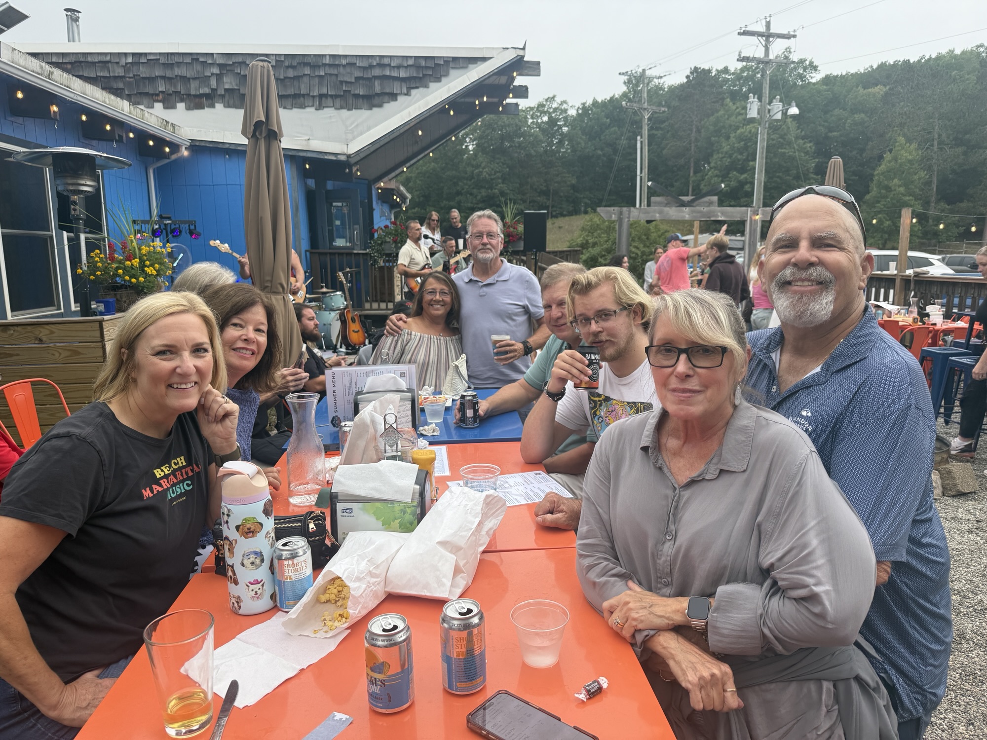 A group of people gathered around a table at an outdoor venue, enjoying drinks and snacks, with live music in the background. The setting features colorful chairs and a blue building adorned with flowers.