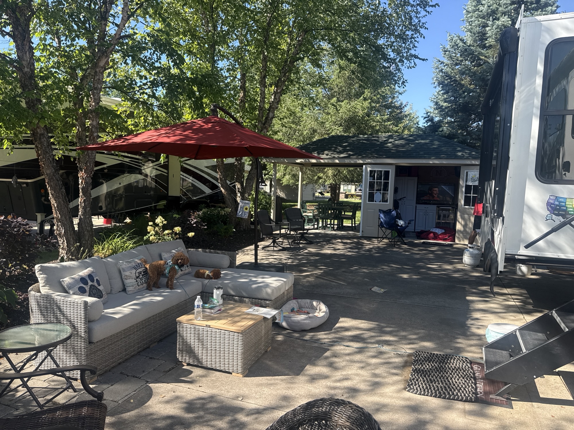 Outdoor seating area with a grey sofa, coffee table, and a red umbrella, surrounded by green trees and an RV in the background.