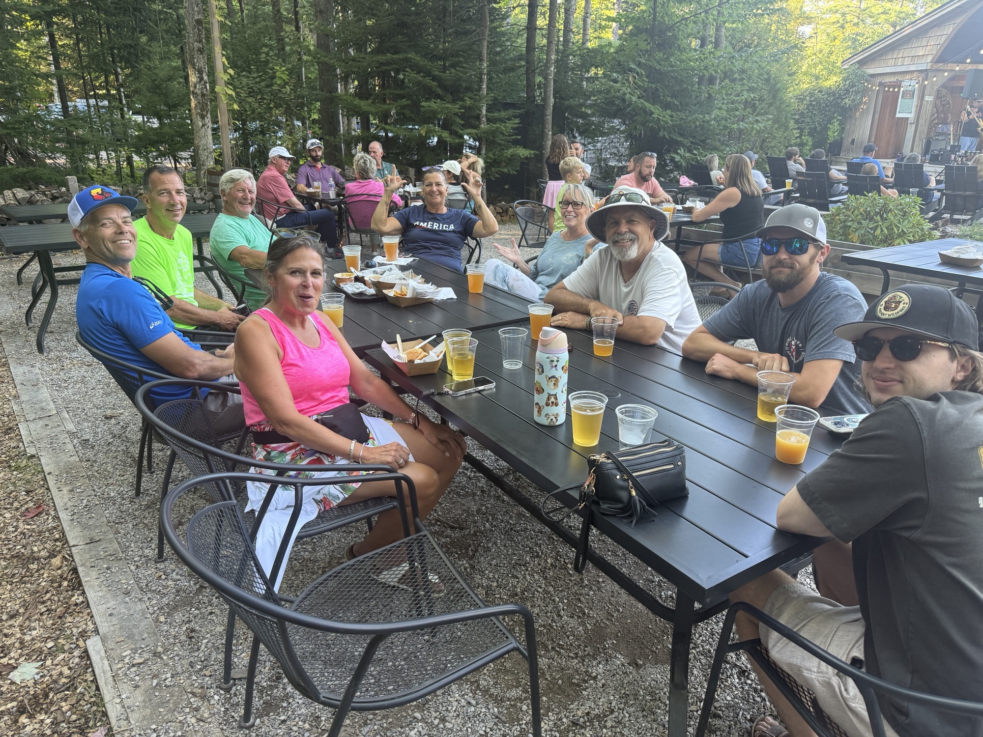 A group of people sitting around a table outdoors, enjoying drinks and food in a forested setting.